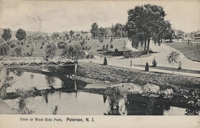 Vintage black and white photograph of West Side Park in Paterson, New Jersey, featuring a small arched bridge over a pond, surrounding trees, a winding path, and park buildings in the background.
