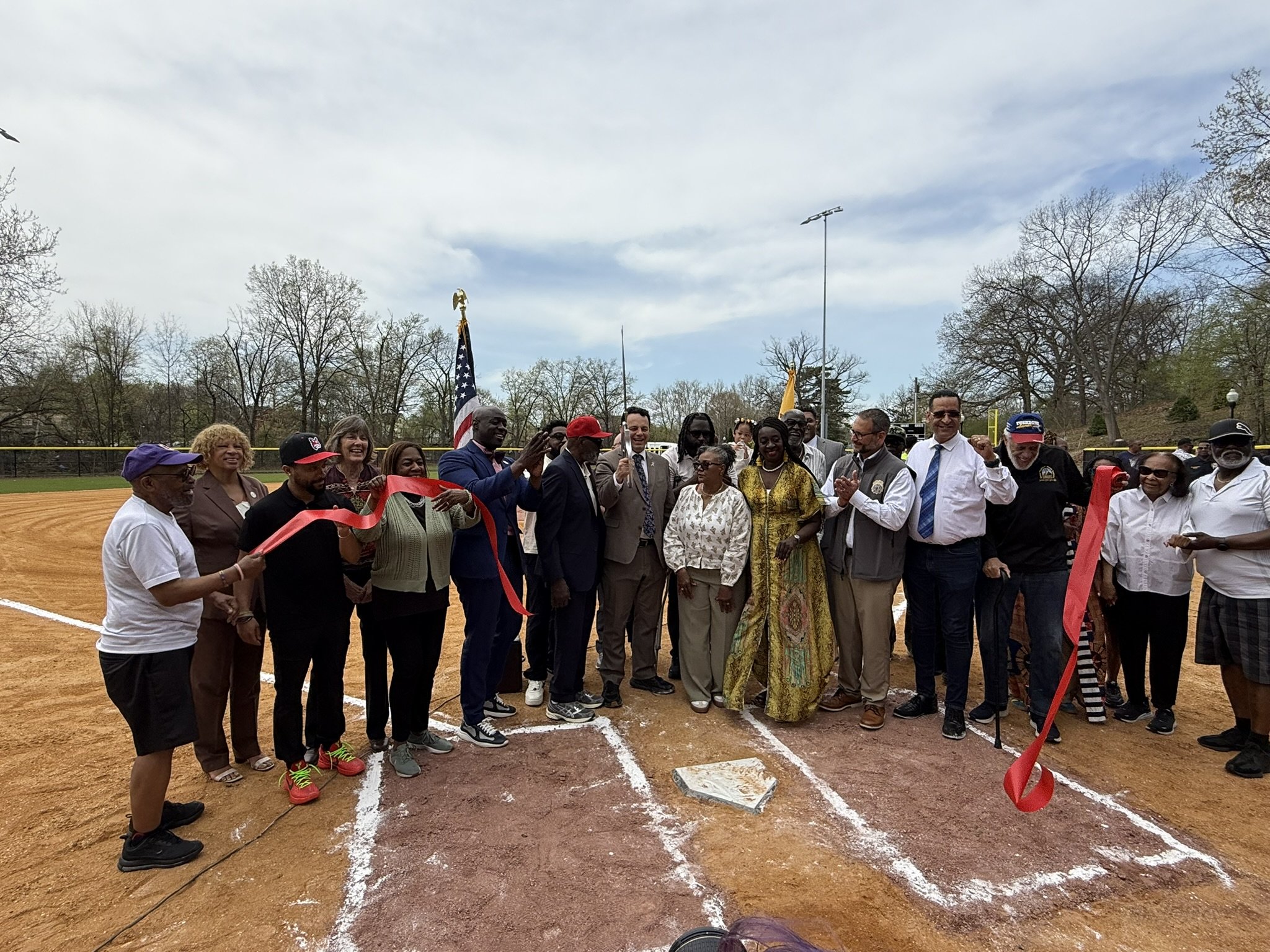A group of diverse people gathered on a baseball field for a ribbon-cutting ceremony, with some holding scissors, an American flag, and smiling.