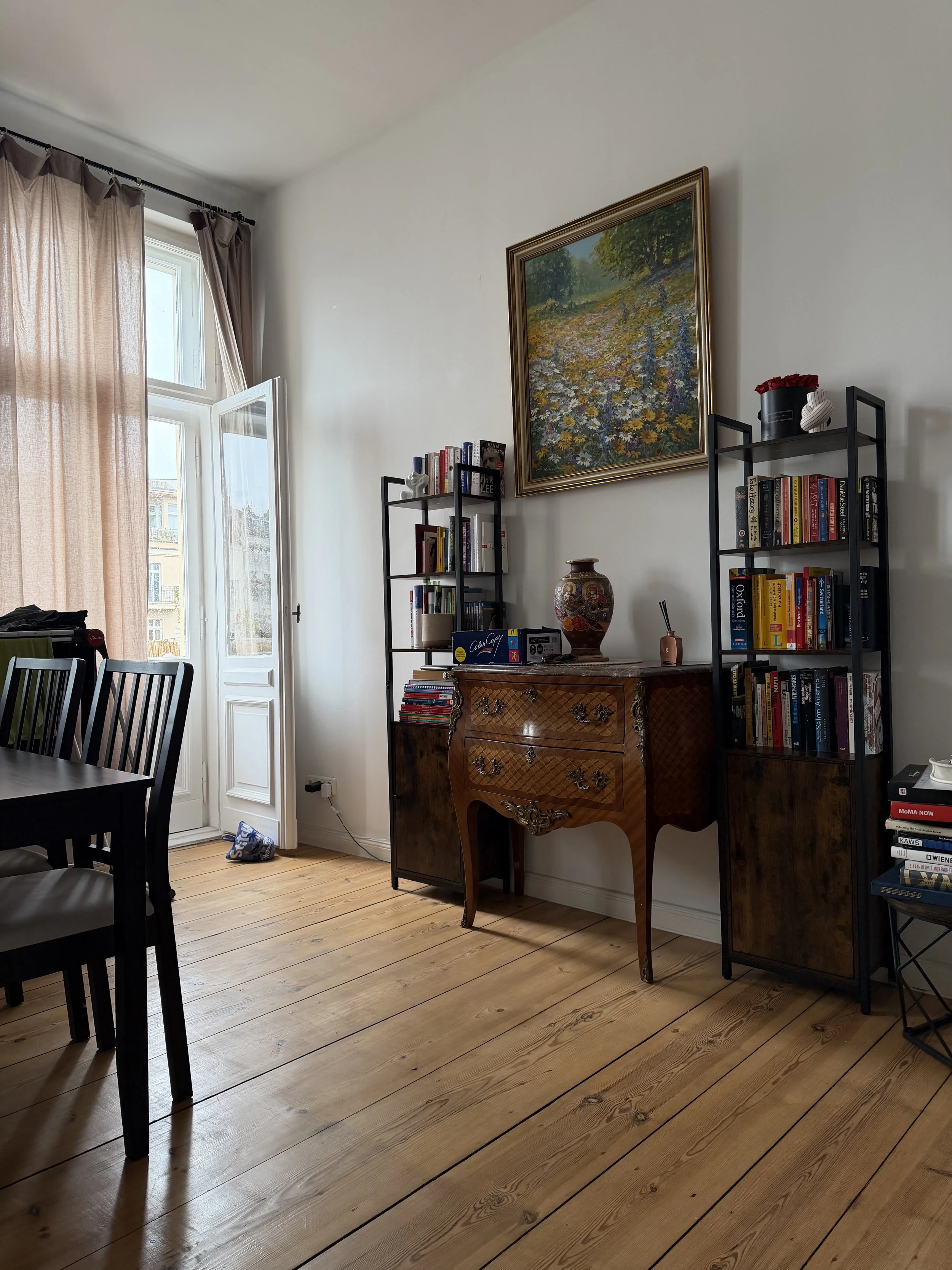 Ein Wohnzimmer mit Holzboden, Bücherregalen, einem großen Gemälde an der Wand und einer offenen Terrassentür, durch die man einen blauen Himmel und Nachbargebäude sieht.