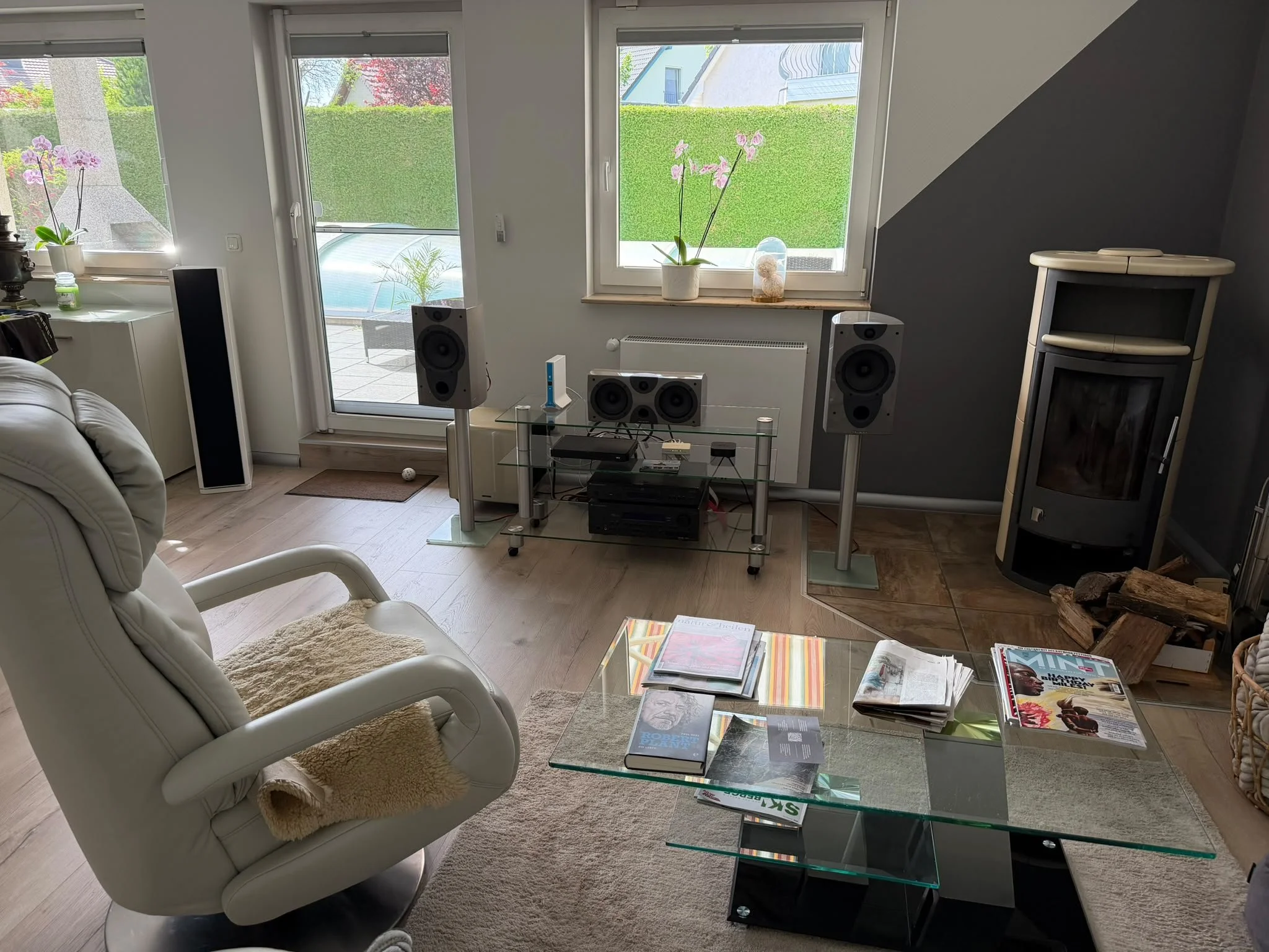 Wohnzimmer mit zwei großen Fenstern und Terrasse im Hintergrund. Es stehen Lautsprecher, ein Fernseher, eine Couch, ein Glastisch mit Magazinen und Büchern, eine Kaminuhr und Dekorationen auf den Fenstersimsen.