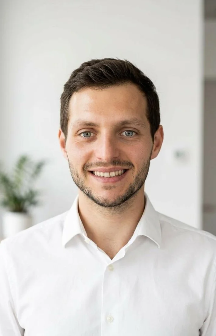 Ein junger Mann mit dunklen Haaren, blauen Augen und einem weißen Hemd lächelt in die Kamera, im Hintergrund ist eine weiße Wand mit einer Topfpflanze sichtbar.