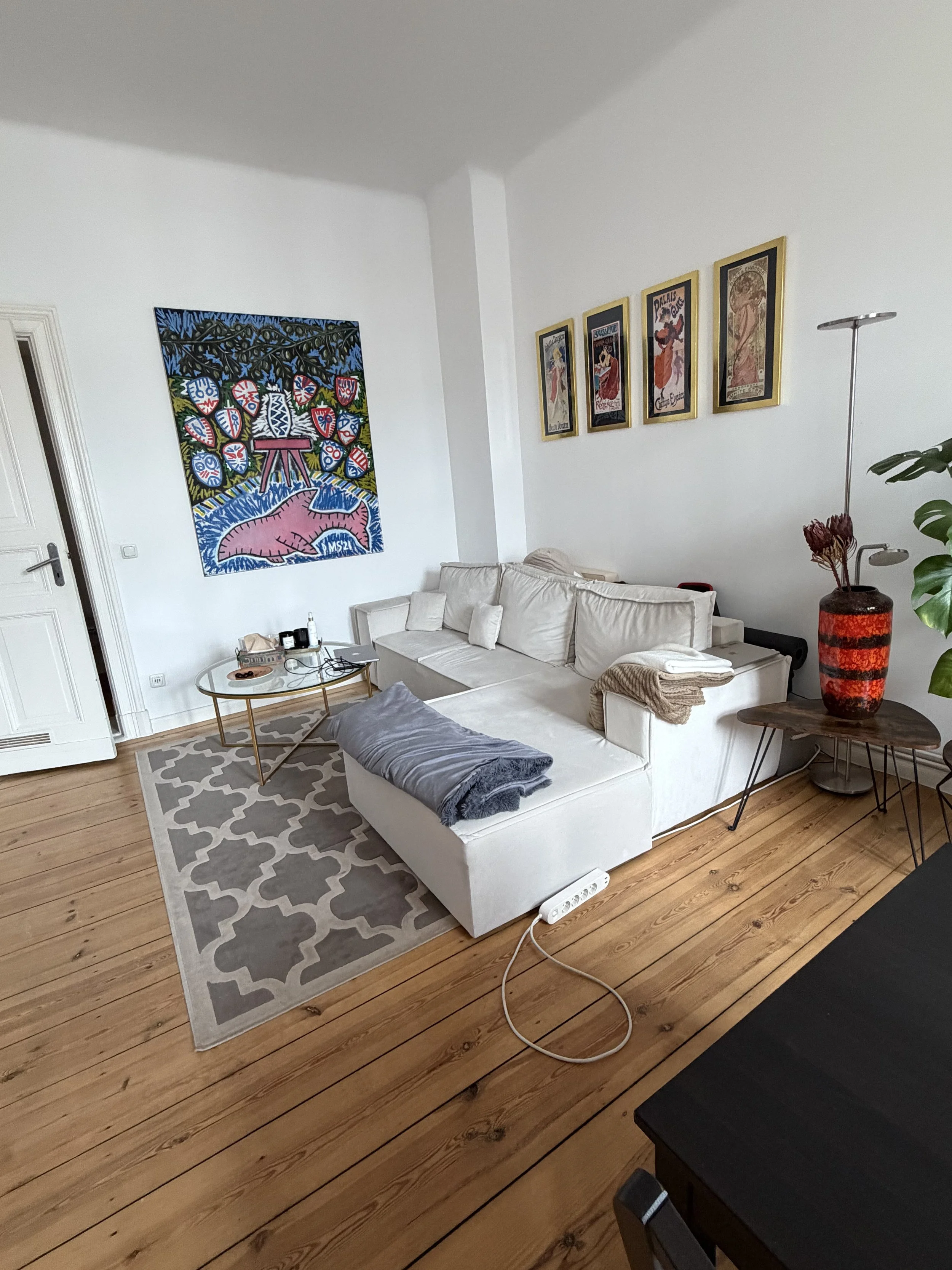Ein Wohnzimmer mit weißem Sofa, einem kleinen runden Couchtisch, Wandkunst mit bunten Gemälden, einem großen roten Vase mit Zweigen, Holzfußboden und einem Teppich.