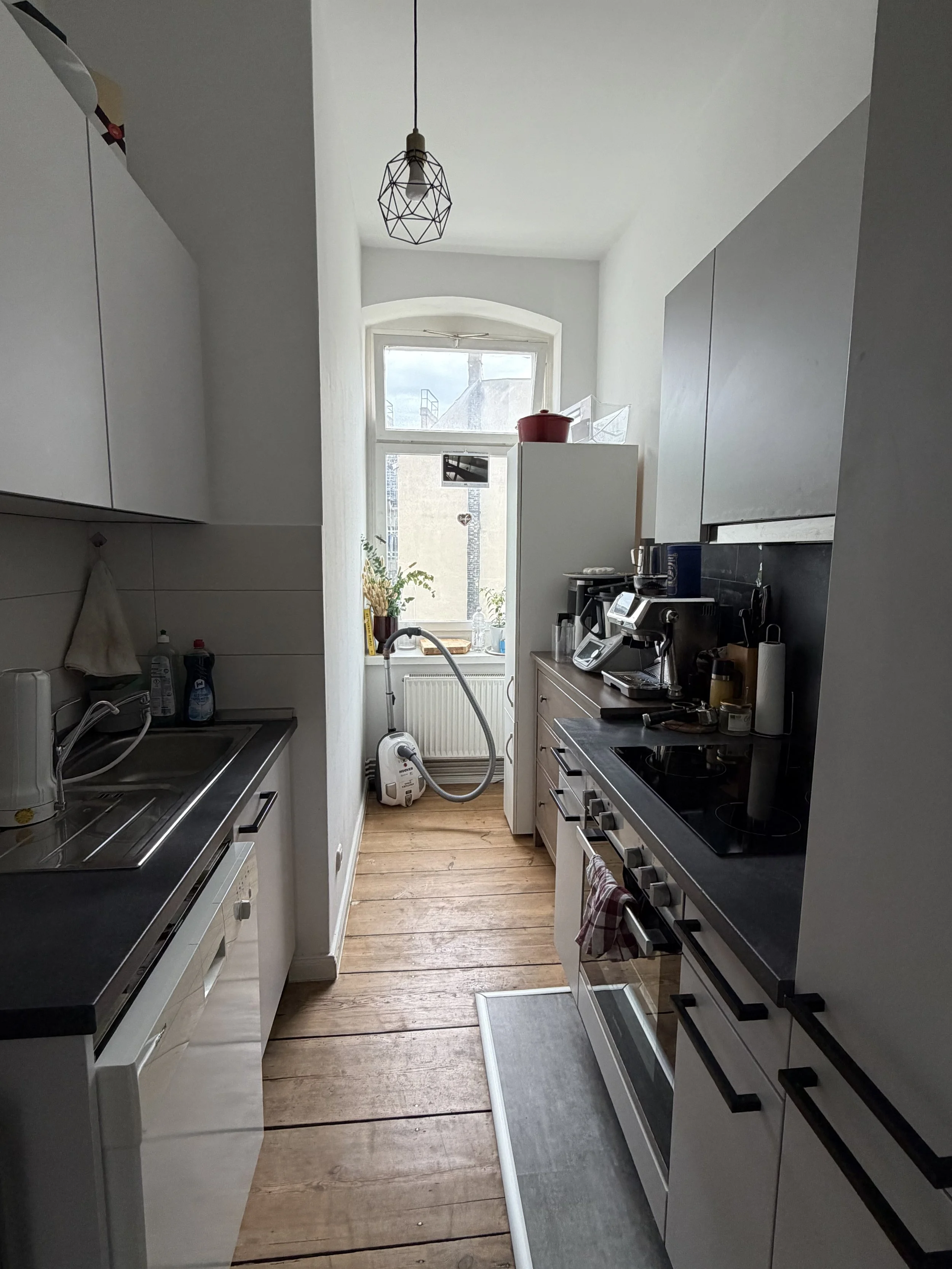 Kleine Küche mit weißen Schränken, schwarzen Arbeitsflächen, Herd, Spüle, Mikrowelle, Kaffeemaschine, Wasserkocher, Pflanzen und einem Fenster mit Blick nach draußen, Holzfußboden.