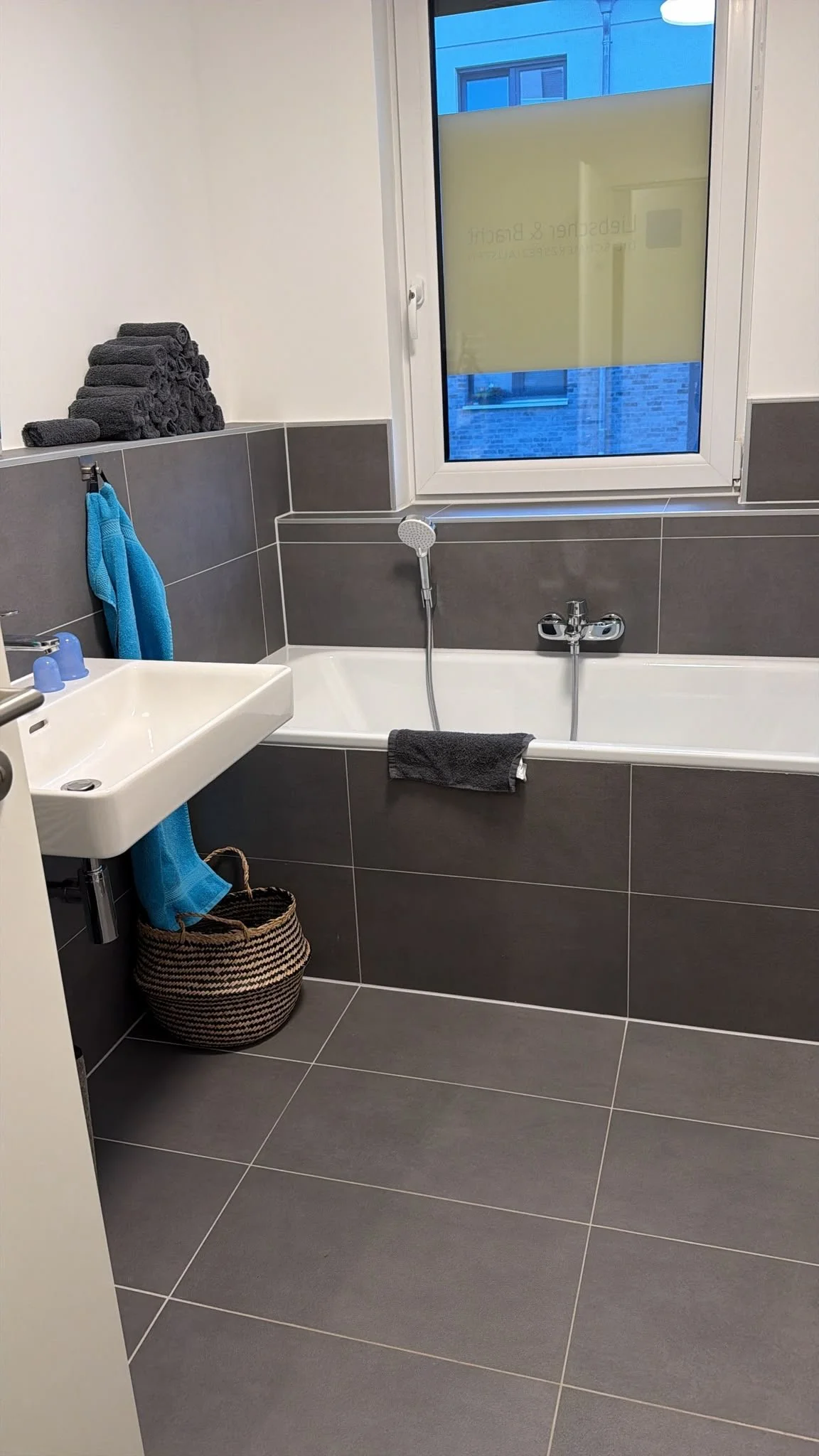 Ein Badezimmer mit grauen Fliesen, einem weißen Waschbecken, einem Badewanne mit Duschkopf, schwarzen und blauen Handtüchern, einem Korb und einem offenen Fenster.