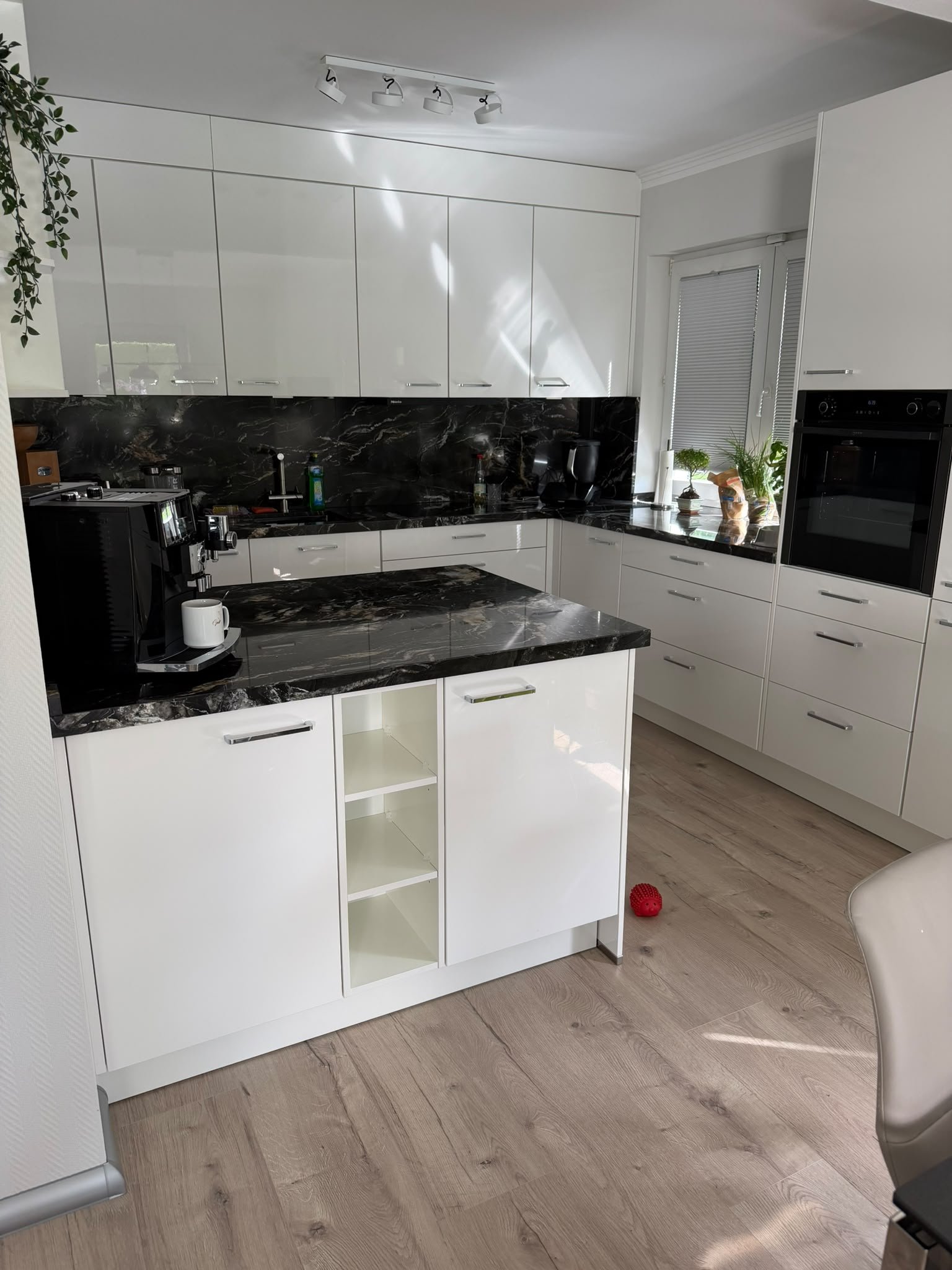 Moderne Küche mit weißen Schränken, schwarzer Marmor-Arbeitsplatte, Kaffeemaschine, Pflanzen auf der Fensterbank und einem roten Spielball auf dem Holzboden.