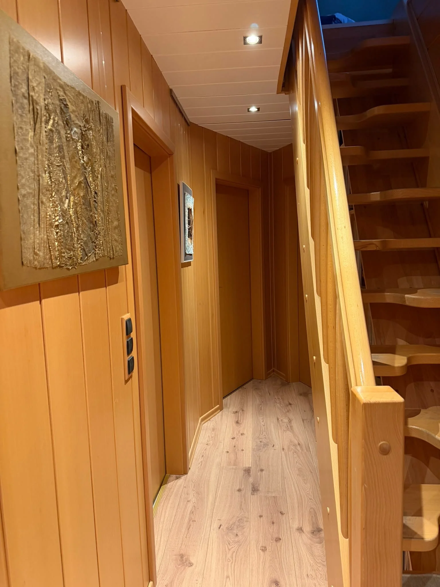 Ein heller Holzboden im Flur eines Hauses mit Holzvertäfelung an den Wänden, einem Geländer und Treppen. An den Wänden hängen zwei Bilder. Die Beleuchtung erfolgt durch in die Decke eingebettete Spots.