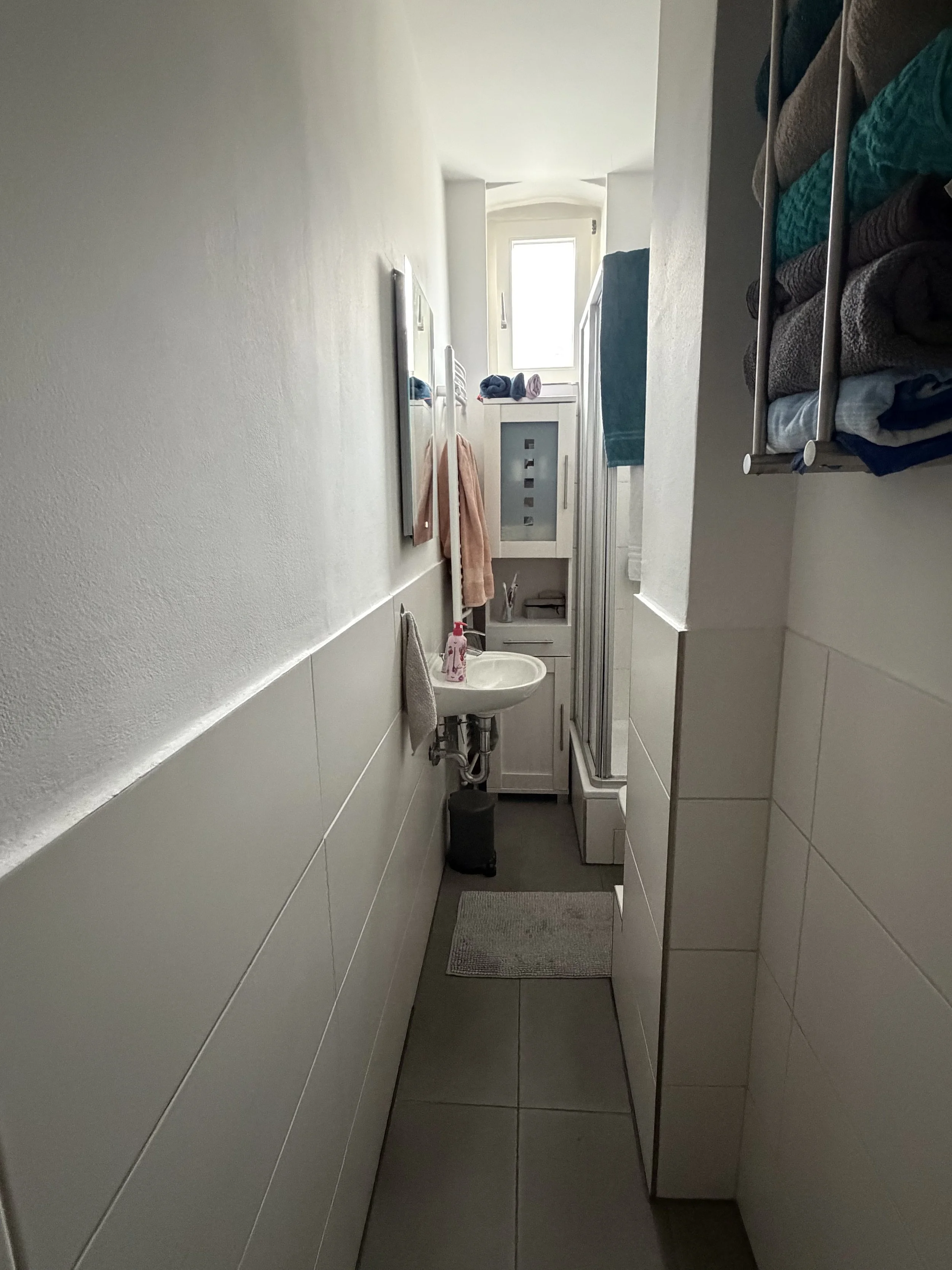 Kleines Badezimmer mit Waschbecken, Spiegel, Handtuch, Dusche und Aufbewahrungsregal mit Handtüchern, Blick zur Fensternear