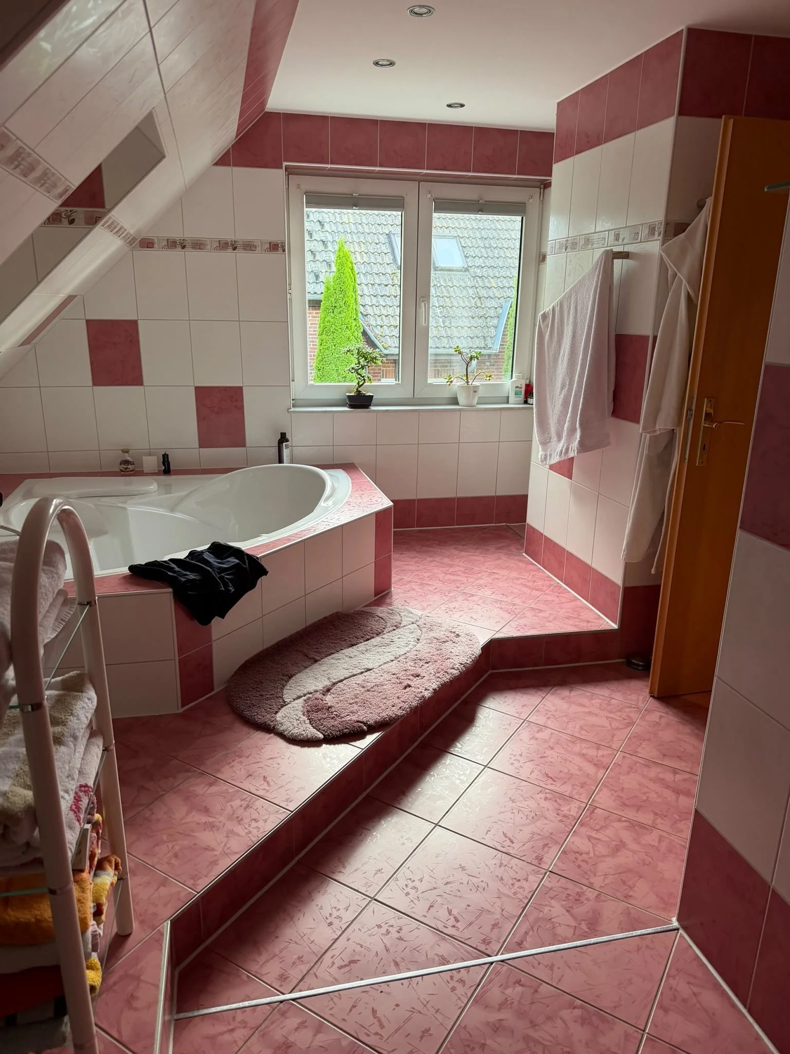 Ein Badezimmer mit Bademantel, Handtüchern und einem Wannenbad, große Fenster mit Blick nach draußen, rote und weiße Wandfliesen, zwei Pflanzen auf dem Fensterbrett, runder Teppich vor der Badewanne, Handtuch an der Wand, eine kleine Flasche auf dem 