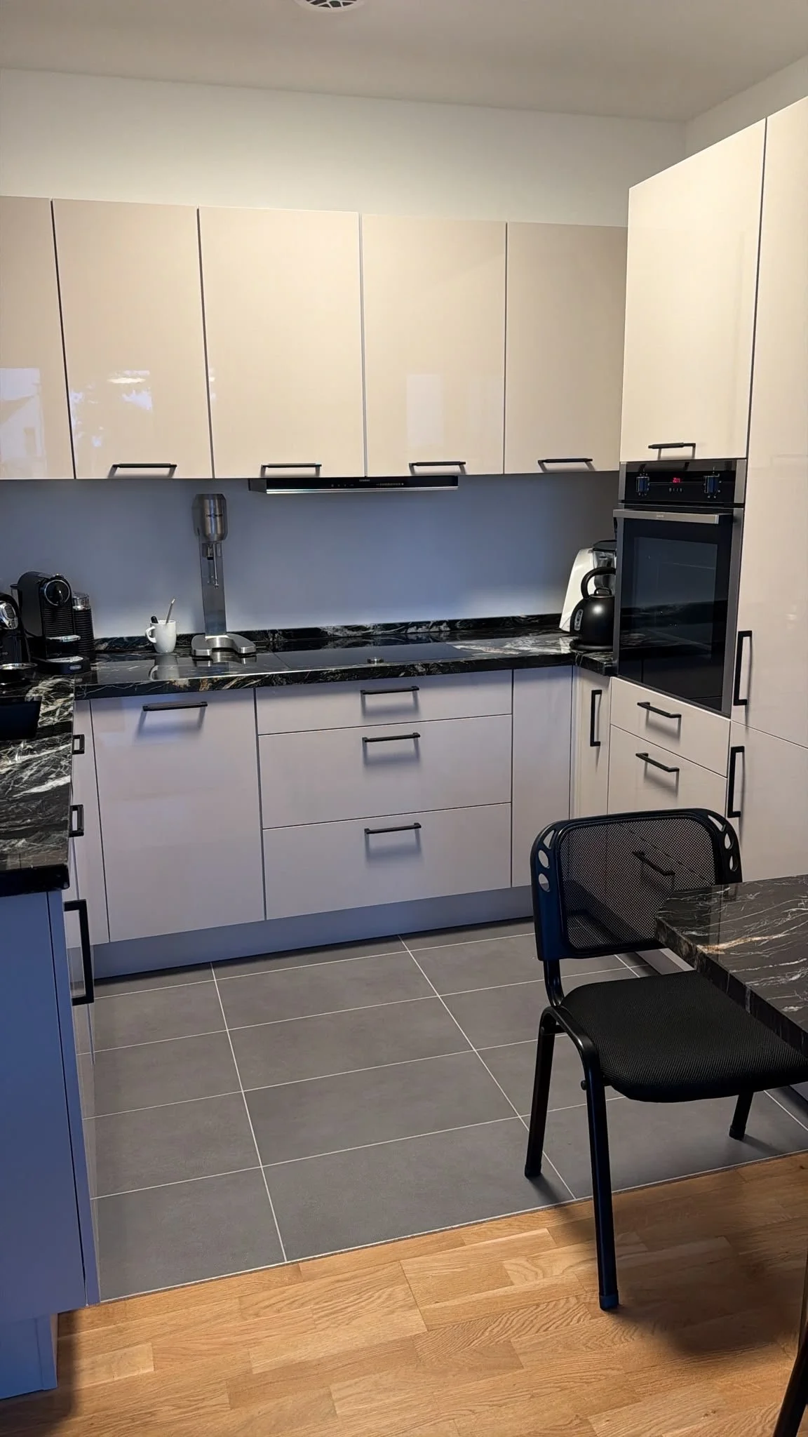 Moderne Küche mit weißen Schränken, schwarzen Arbeitsplatten, einem eingebauten Ofen, Kaffeemaschine, Wasserkocher und Sitzgelegenheit.