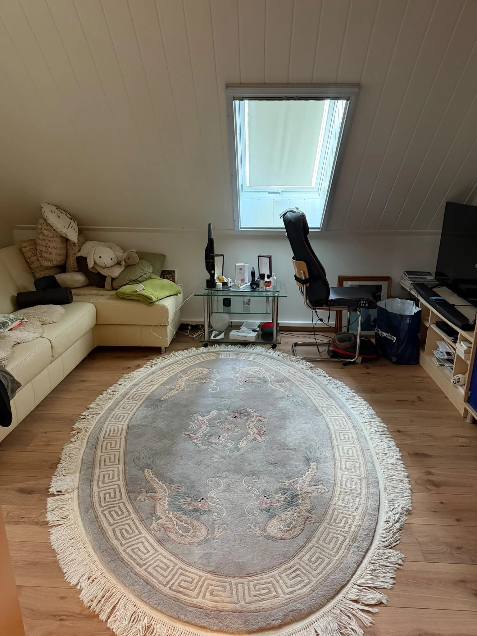 Ein Wohnzimmer mit einem runden, beigefarbenen Teppich mit chinesischen Drachenmustern, einem weißen Sofa mit Kissen und Plüschspielzeug, einem Schreibtisch mit Gegenständen, einem Fernseher und einem Fenster im Dach mit geschlossenem Rollladen.