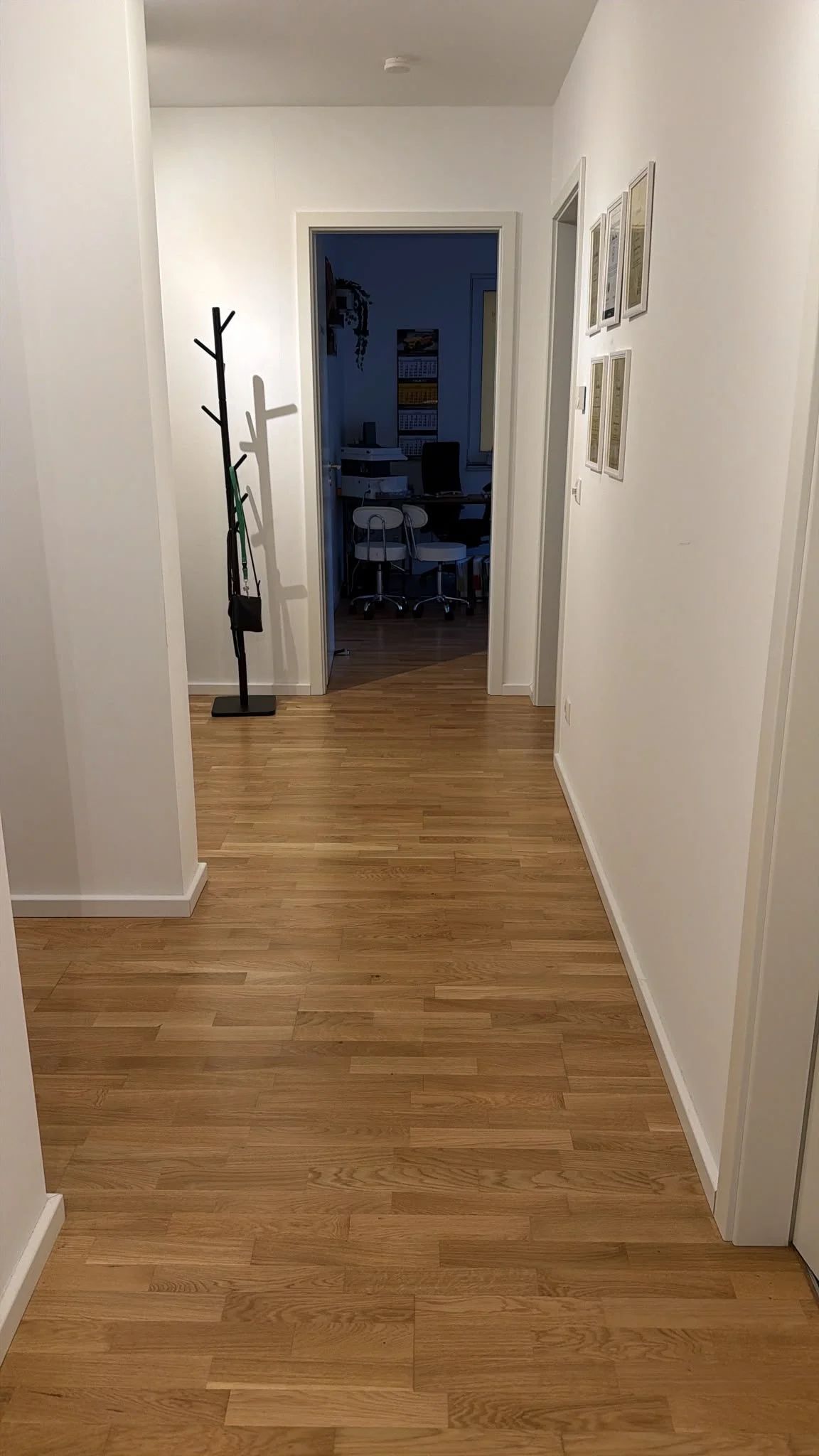 Der Flur in einem Haus mit hellem Holzboden, weißen Wänden und mehreren Bildern an der rechten Wand. Am Ende des Flurs ist ein Raum mit einem Schreibtisch, einem Bürostuhl und einem roten Stuhl. An der linken Seite steht ein schwarzer Garderobenständ