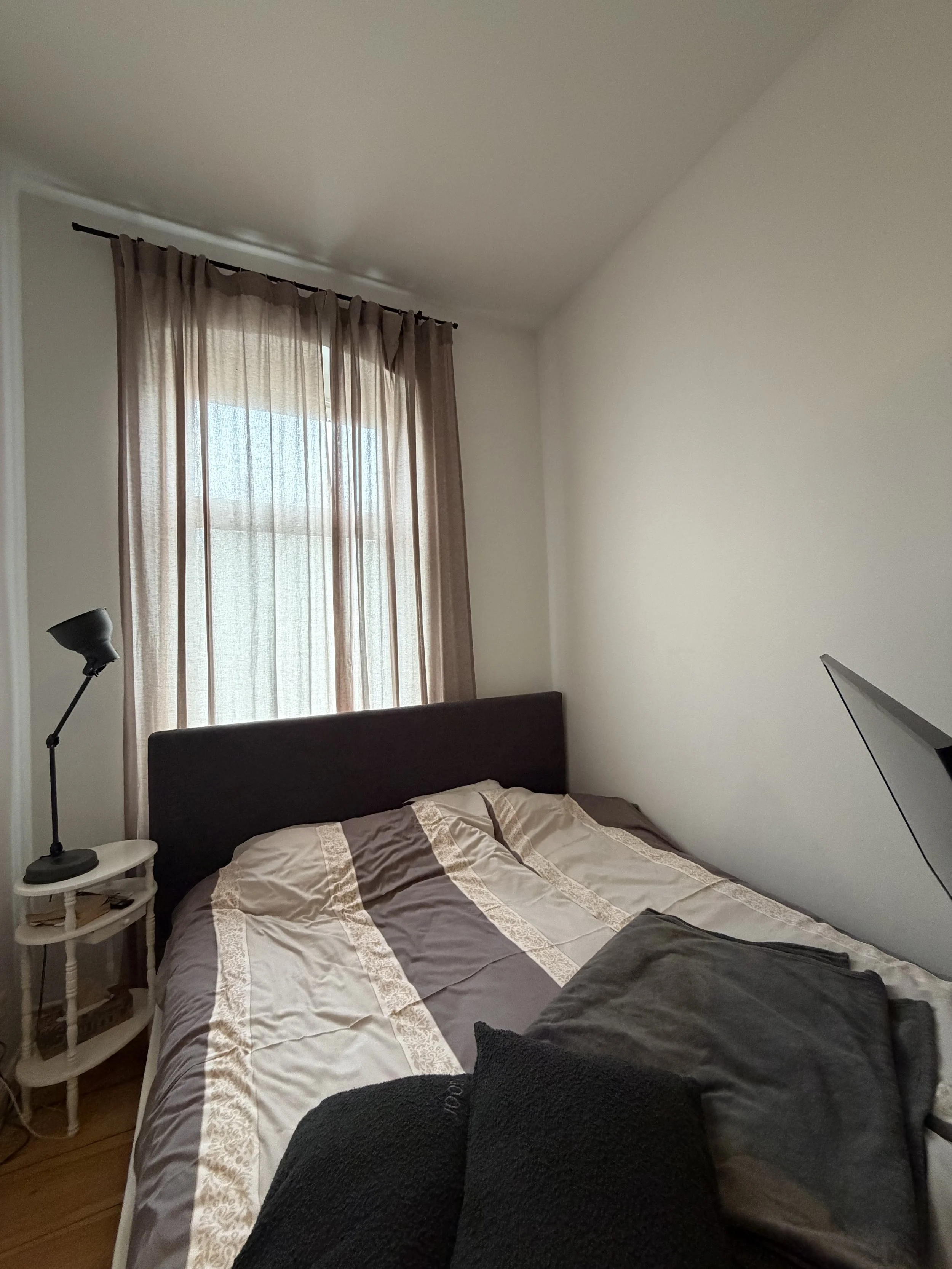 Ein Schlafzimmer mit einem großen Bett mit beige und brauner Bettwäsche, einem Fenster mit hellbraunen Vorhängen, einem Nachttisch mit einer Lampe und einem Buch, und einem Fernseher auf einem Tisch.
