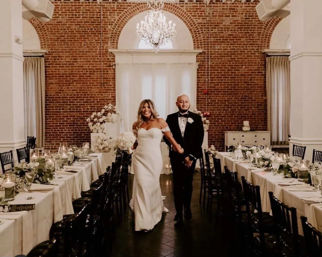 Bride and groom holding hands inside a decorated wedding reception hall with brick walls, chandelier, and banquet tables with floral centerpieces.