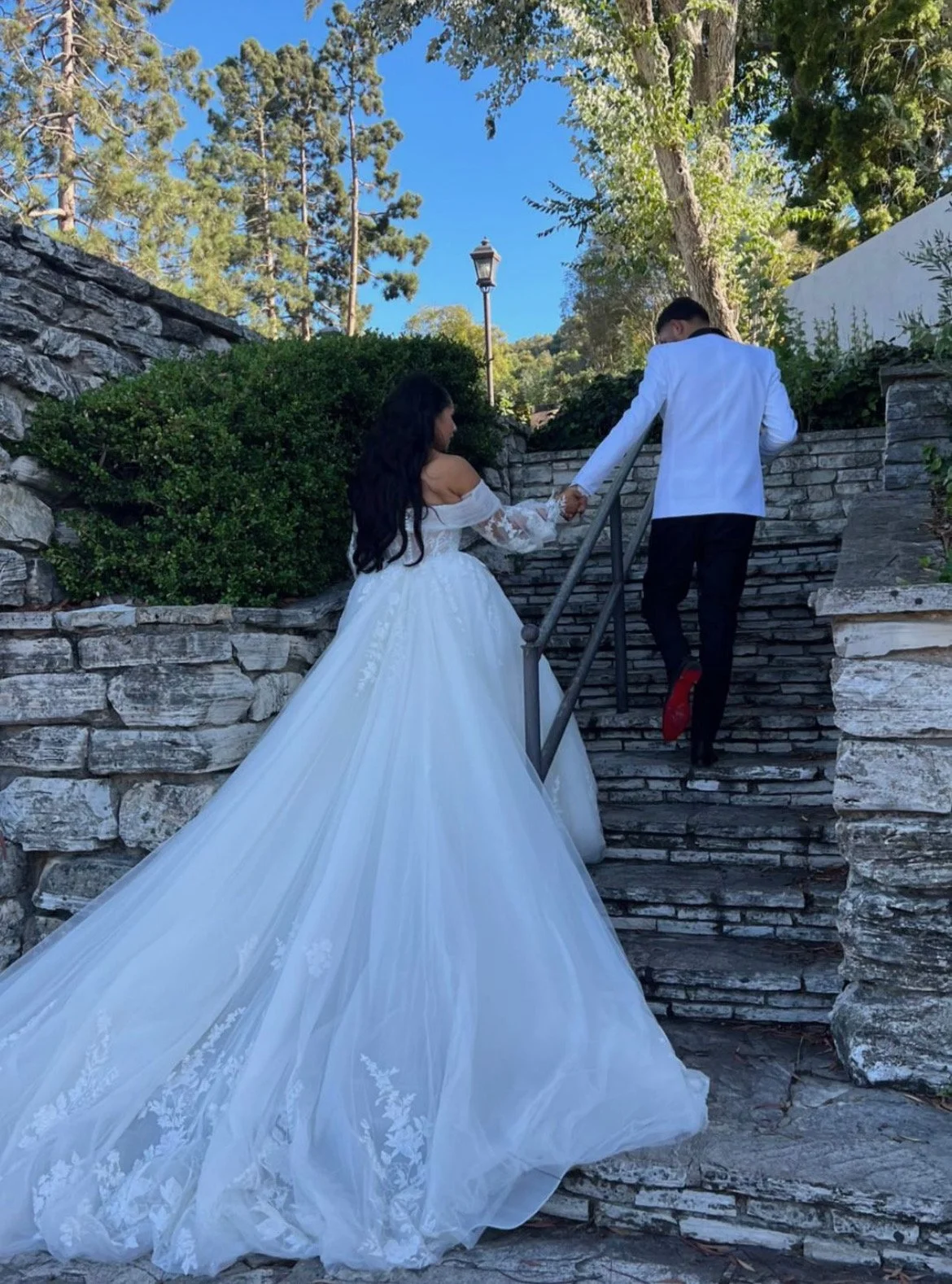 A bride and groom holding hands while climbing stone stairs outdoors, with the bride in a white wedding gown and the groom in a white jacket and black pants.