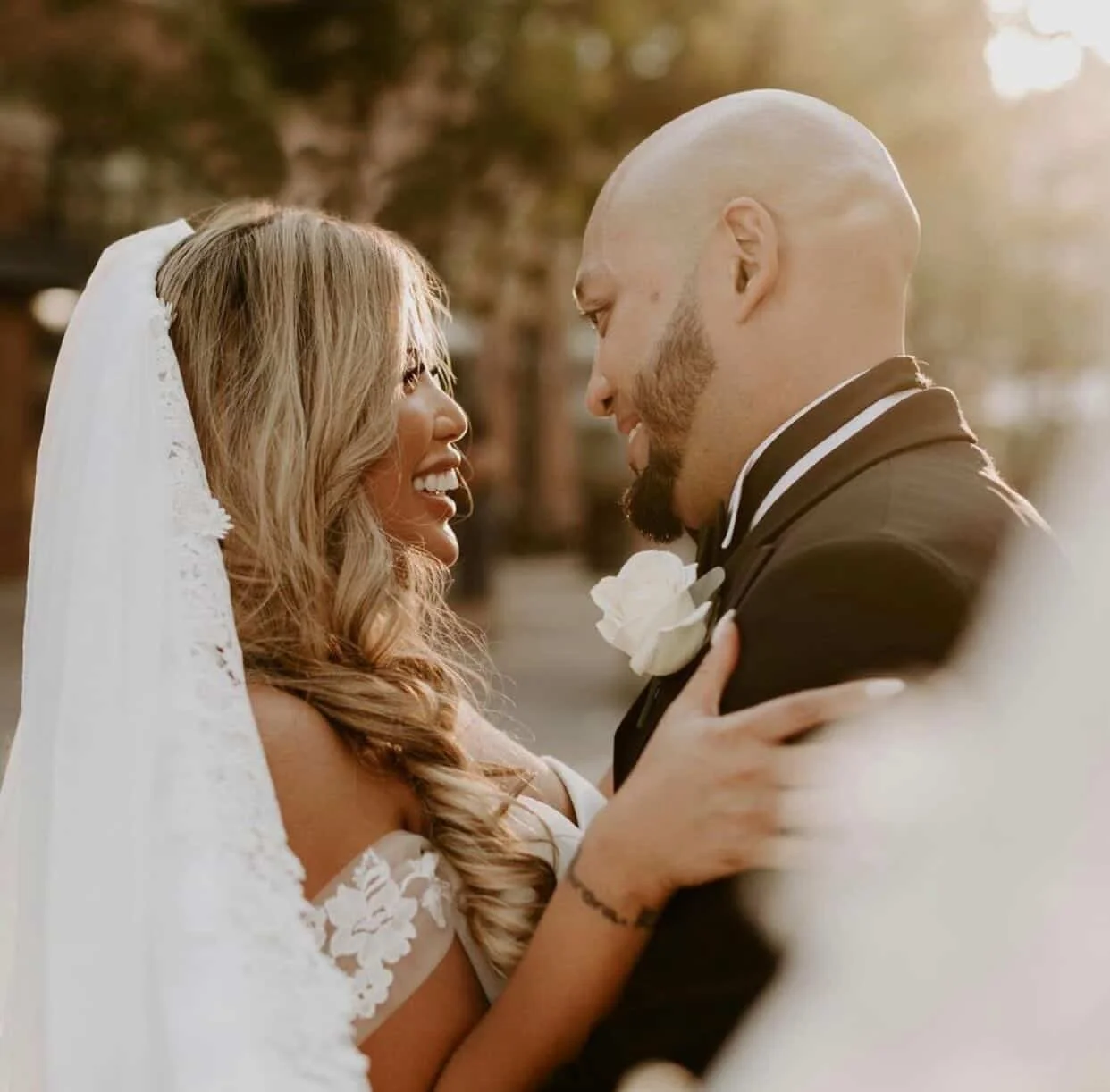 A bride and groom looking at each other outdoors during sunset, sharing an intimate moment, with the bride wearing a wedding dress and veil, and the groom in a tuxedo with a white boutonniere.