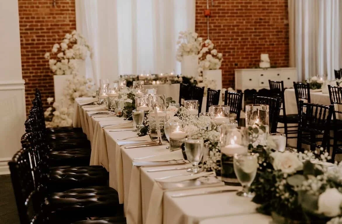 Elegant banquet table decor with white floral arrangements, candles, and glassware in a warmly lit event space.
