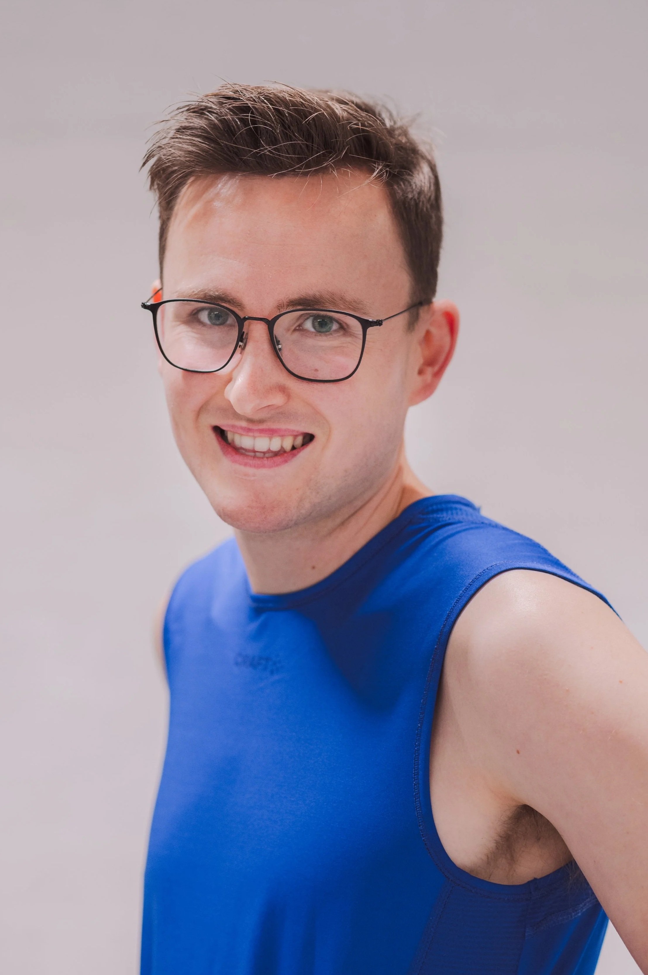 A young man with short brown hair, glasses, and a smile wearing a blue sleeveless athletic shirt, taking a selfie against a plain white background.