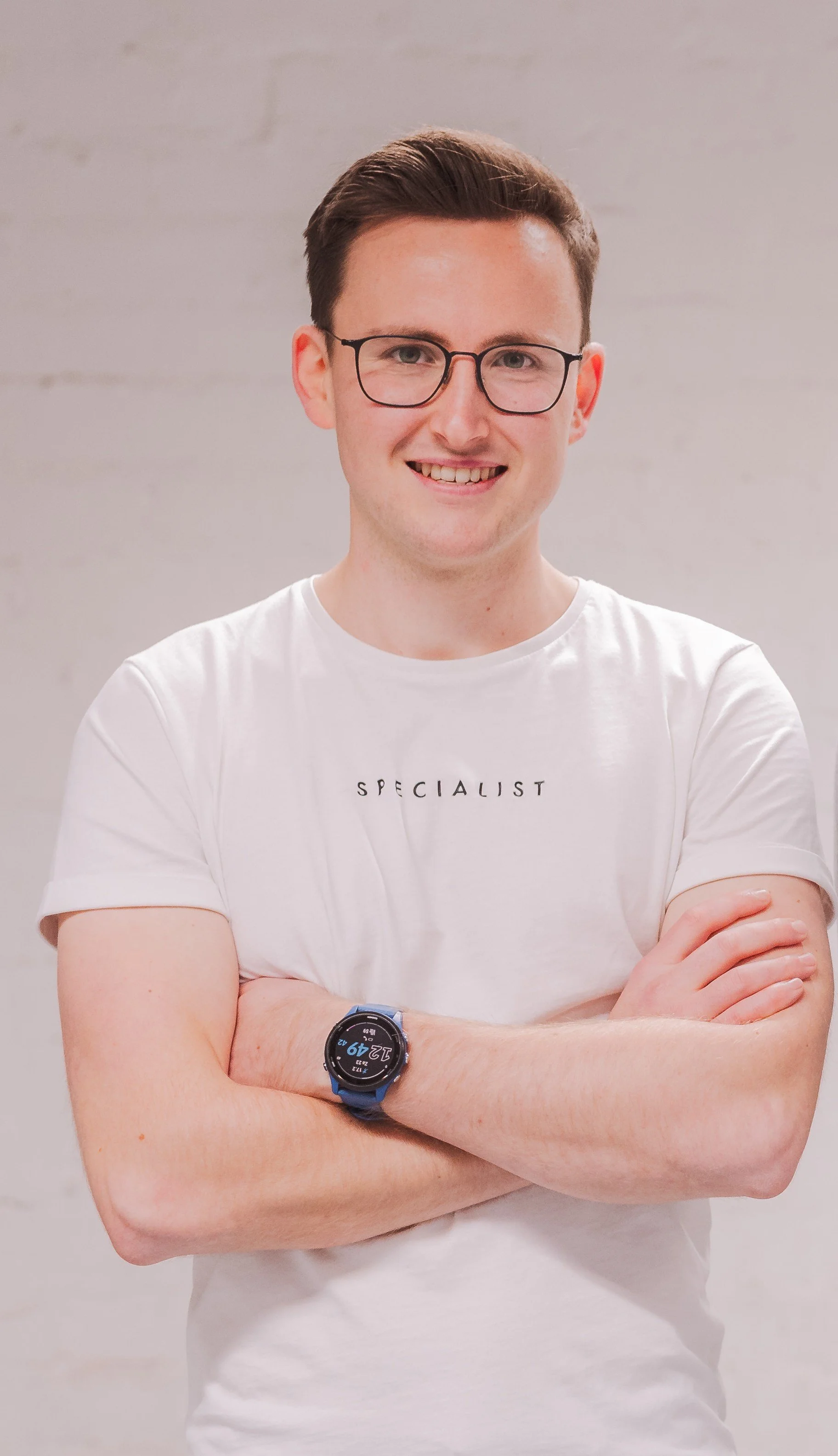 A young man with glasses smiling, arms crossed, wearing a white T-shirt with the word 'SPECIALIST' on it, and a sports watch on his left wrist, standing against a plain light-colored wall.
