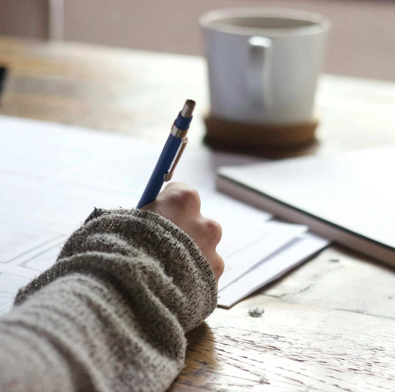 A person's hand holding a blue pen writing on a piece of paper with a small pile of books in the background and a white mug on a table.