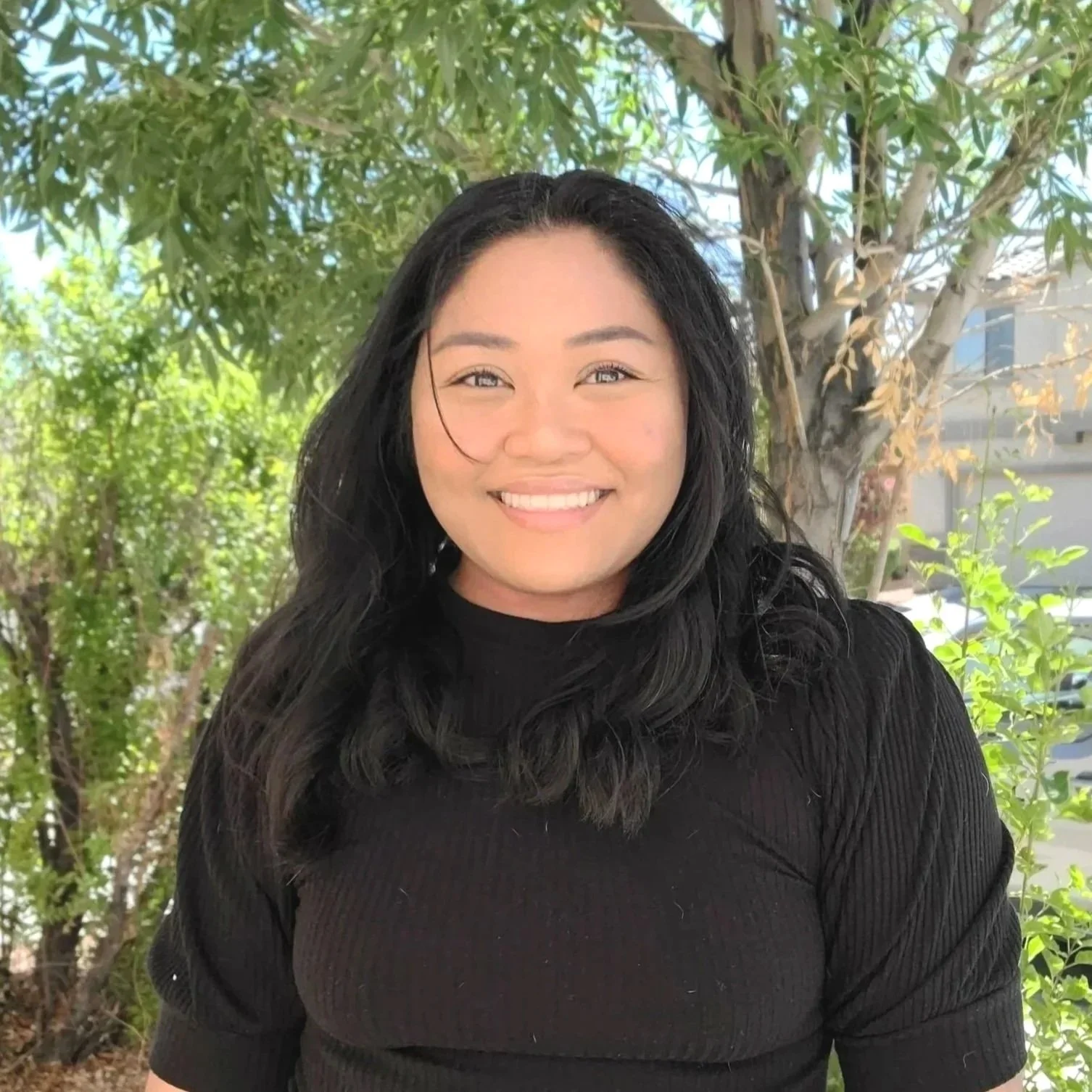 A smiling woman with long dark hair in an outdoor setting with green trees and foliage behind her.