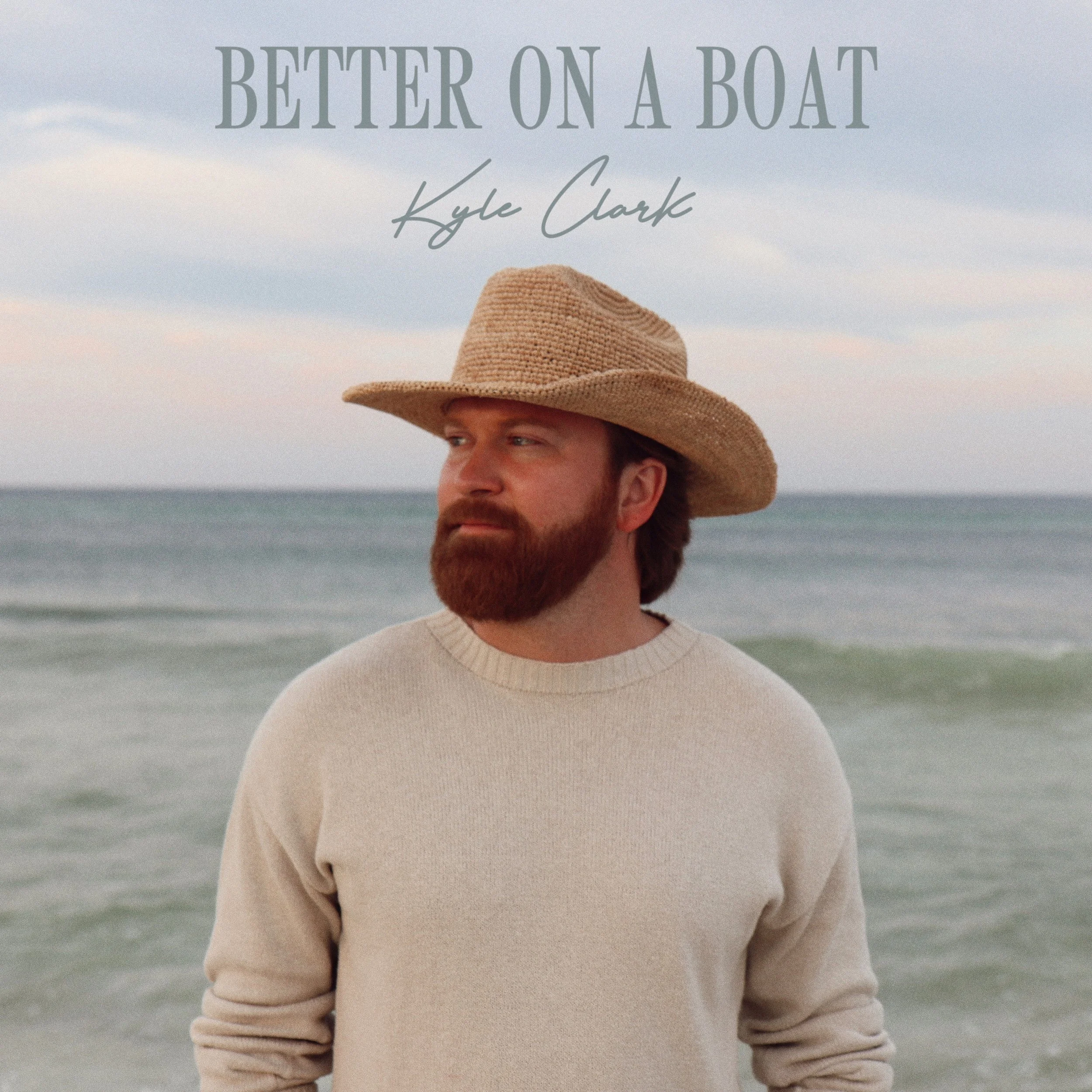 Close-up of a man with a red beard wearing a wide-brimmed straw hat, standing on the beach with ocean waves in the background.