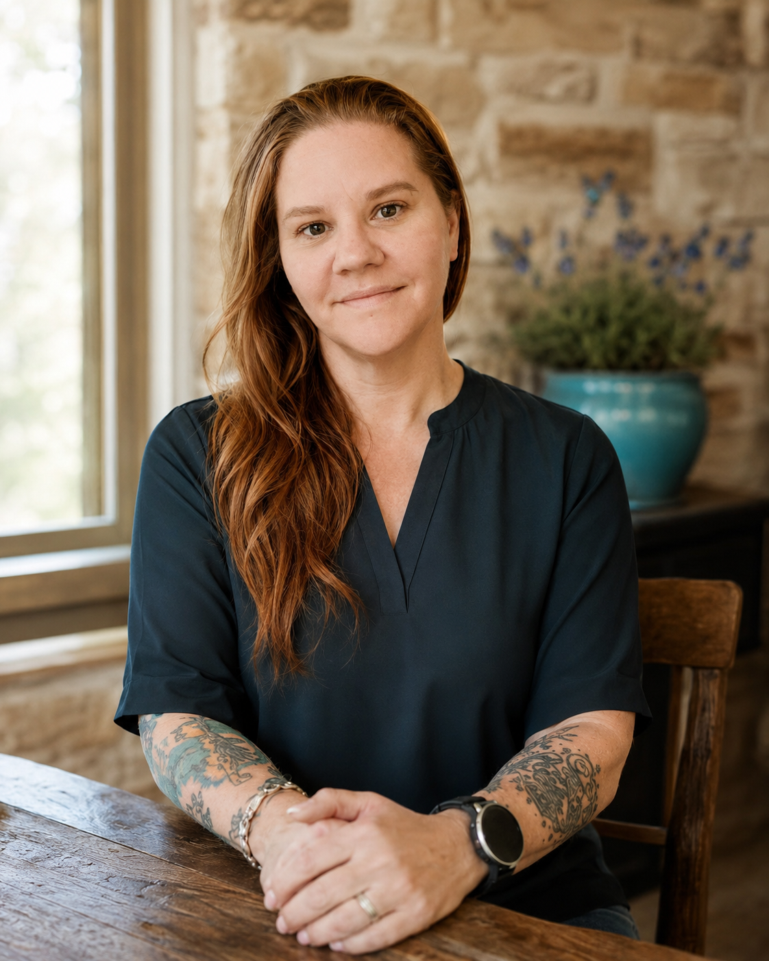 A woman with long red hair wearing a dark blue top, sitting at a wooden table with her hands folded, indoors near a window with a brick wall and a potted plant in the background.