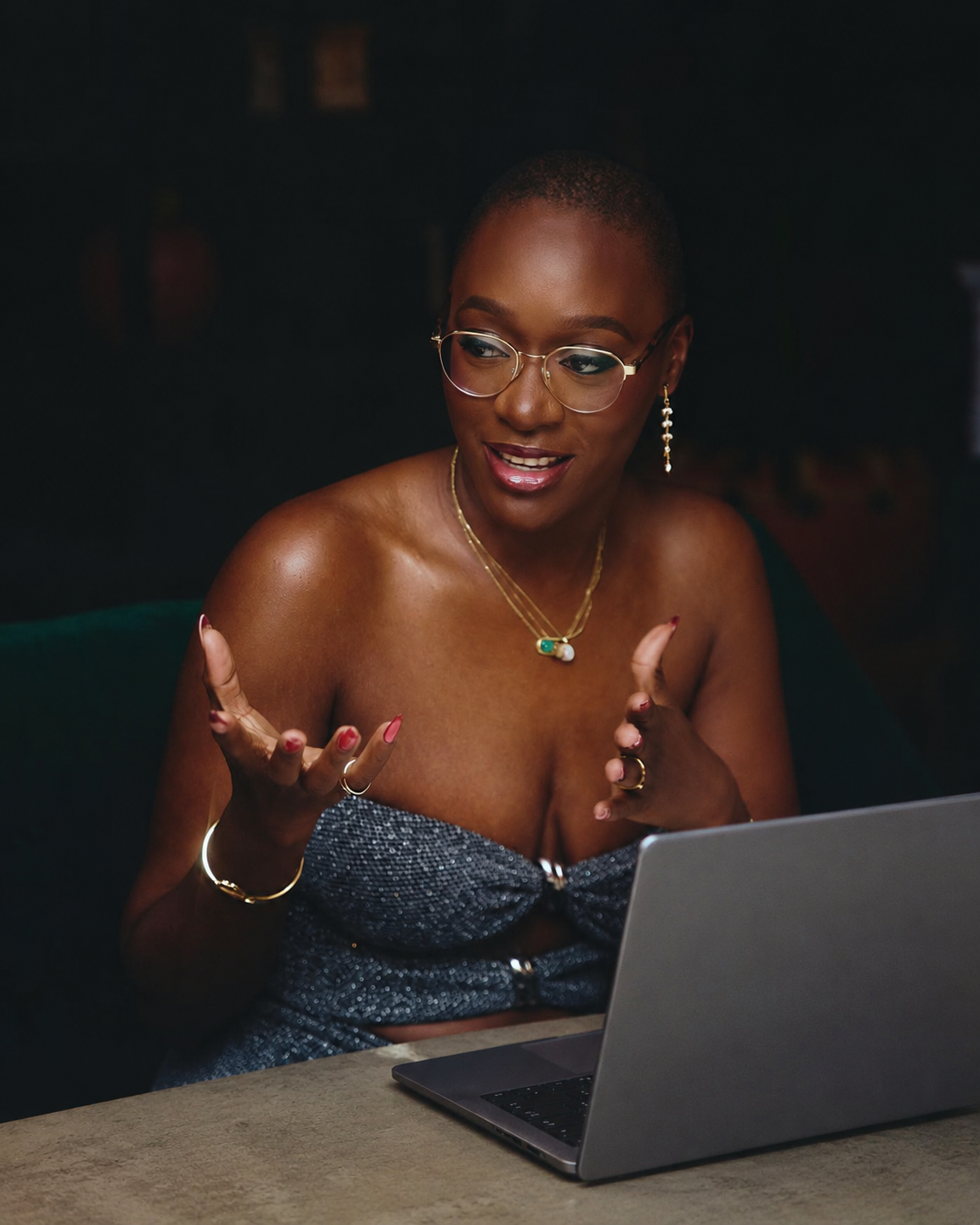 A woman with glasses, short hair, and jewelry is sitting at a table in front of a laptop, gesturing with her hands and speaking.