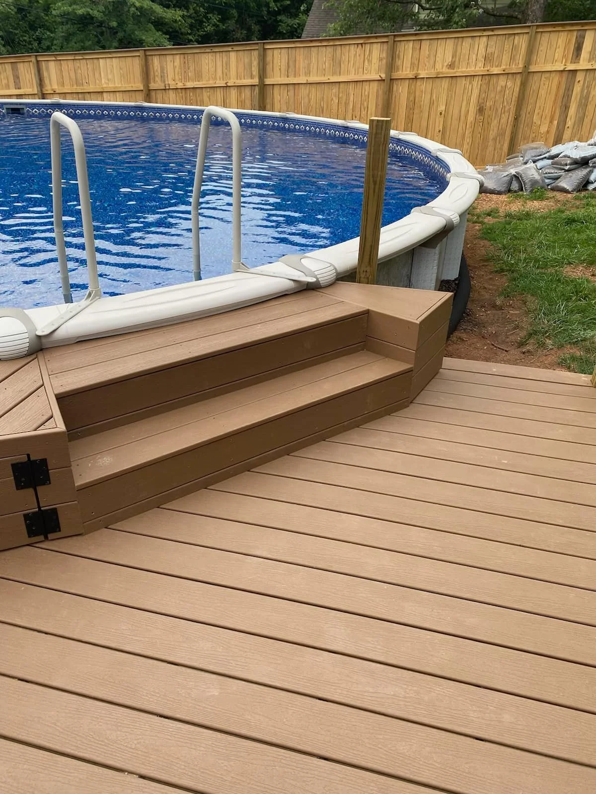 An above view of a backyard swimming pool with wooden steps leading into the water, surrounded by a wooden deck and a yard with grass and a wooden fence.