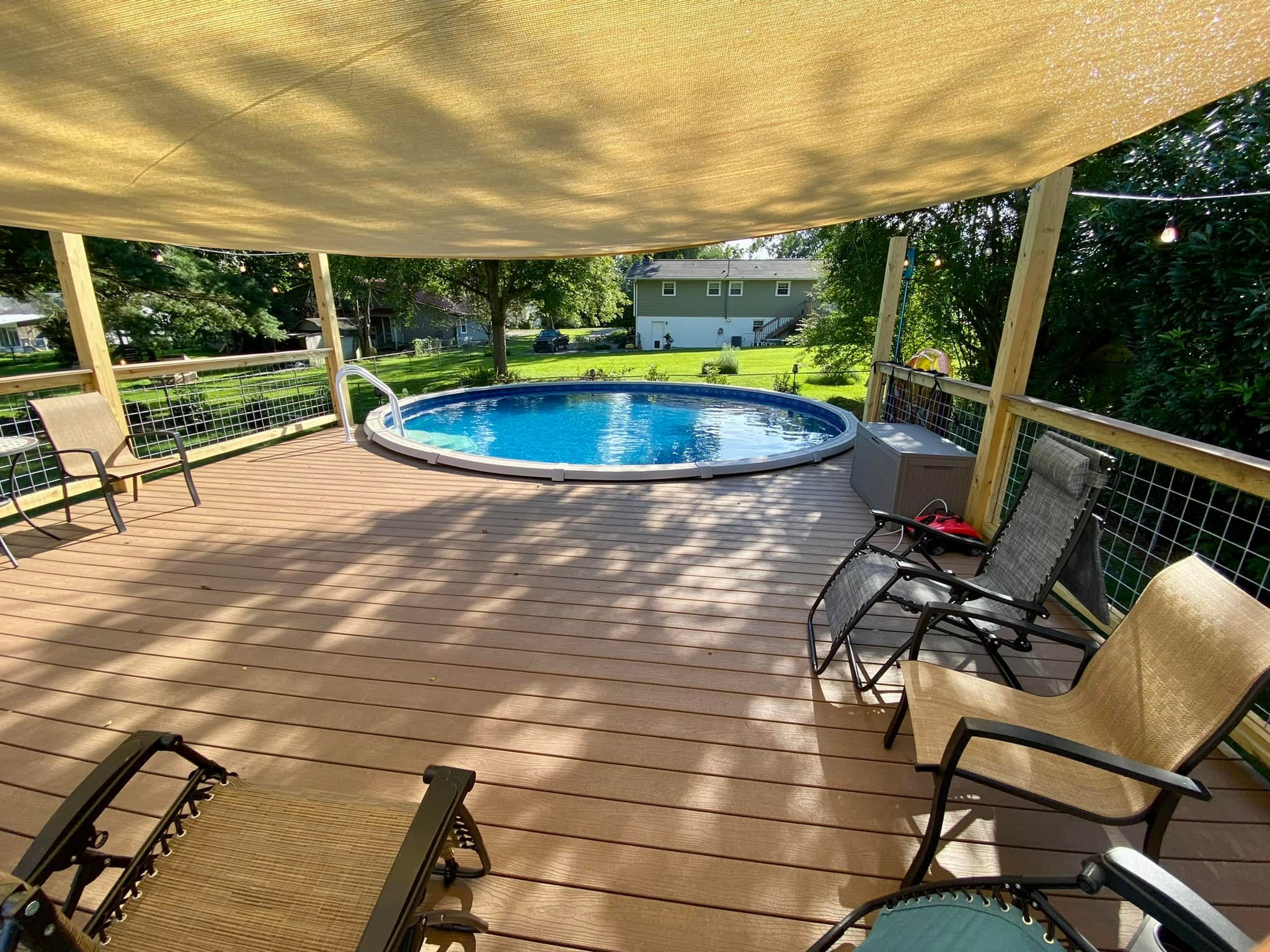 An outdoor backyard deck with a small round above-ground swimming pool, several chairs, and a shaded sun canopy overhead. The background features a green lawn, trees, and a house.