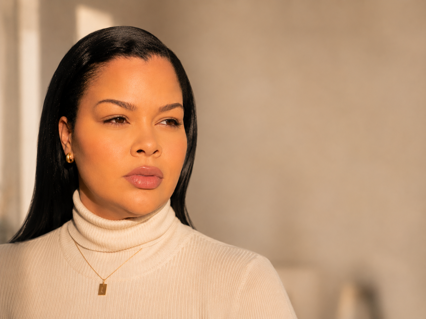 Close-up of a woman with black hair, wearing a beige turtleneck, gold earrings, and a gold necklace with a square pendant, looking to her left with a neutral expression.