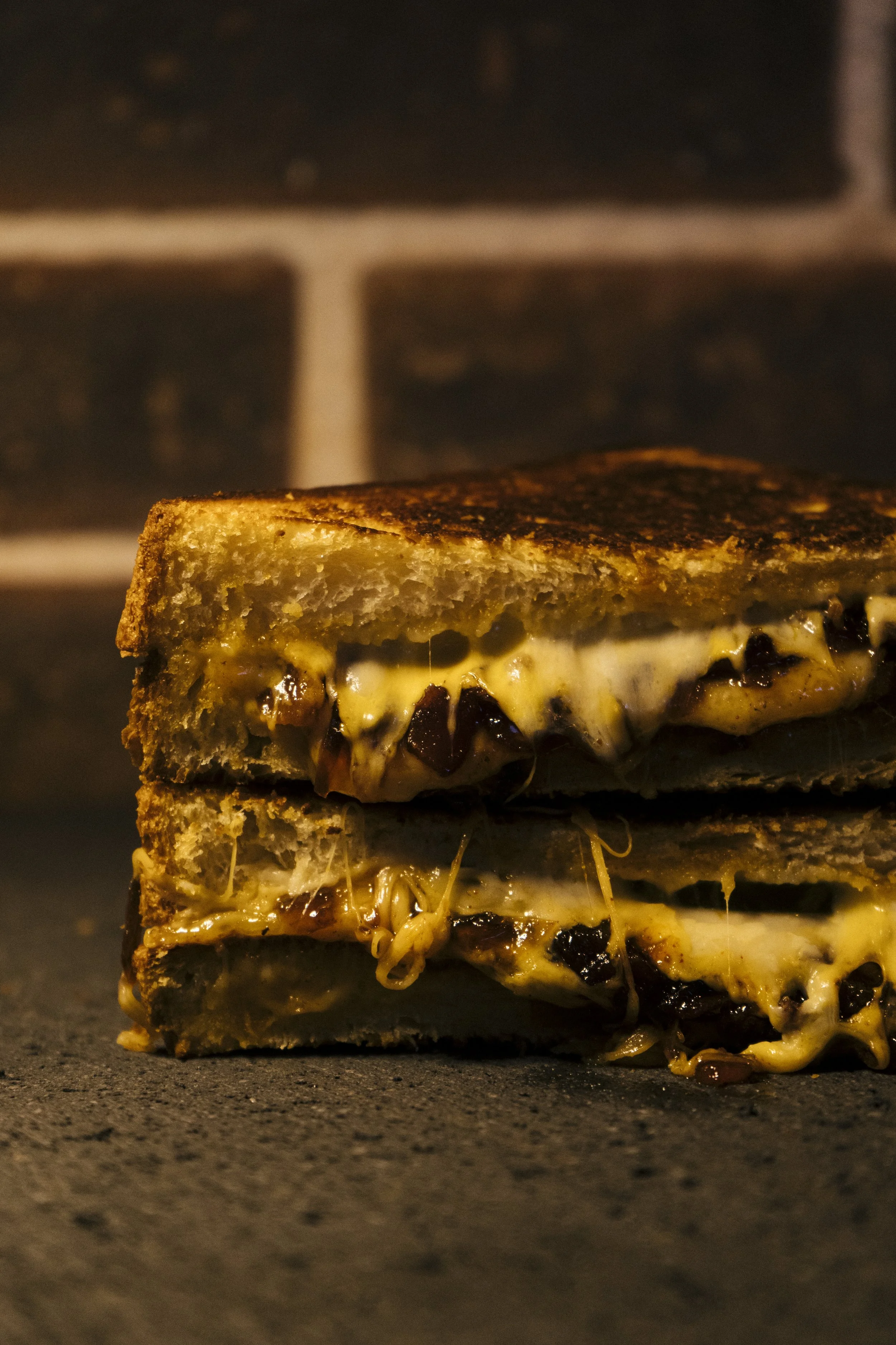 Close-up of a grilled sandwich with melted cheese, dark sauce, and sprouts, resting on a dark surface with a blurred oven in the background.