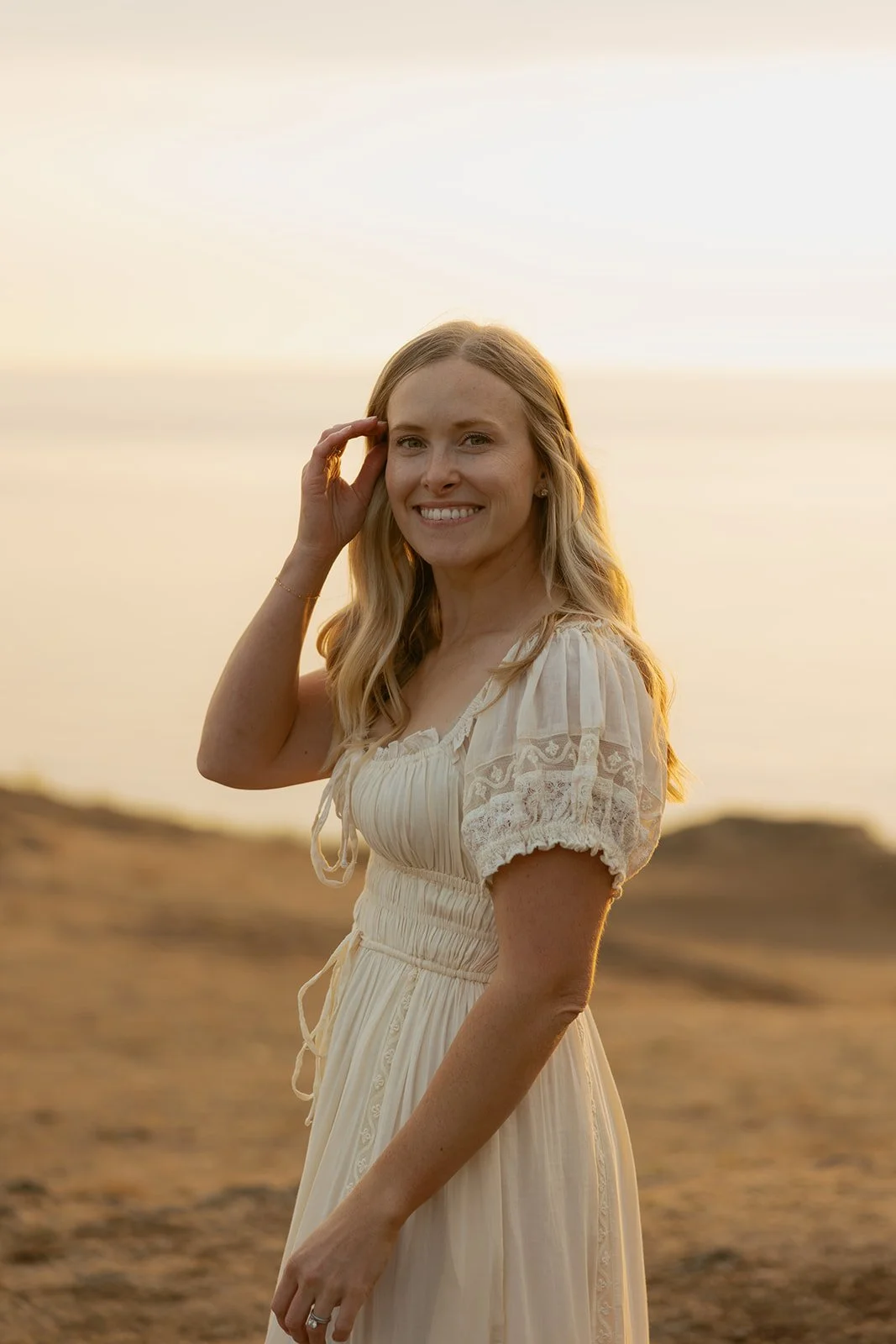 A woman smiling at the camera on a beach during sunset, wearing a light-colored dress with lace details.