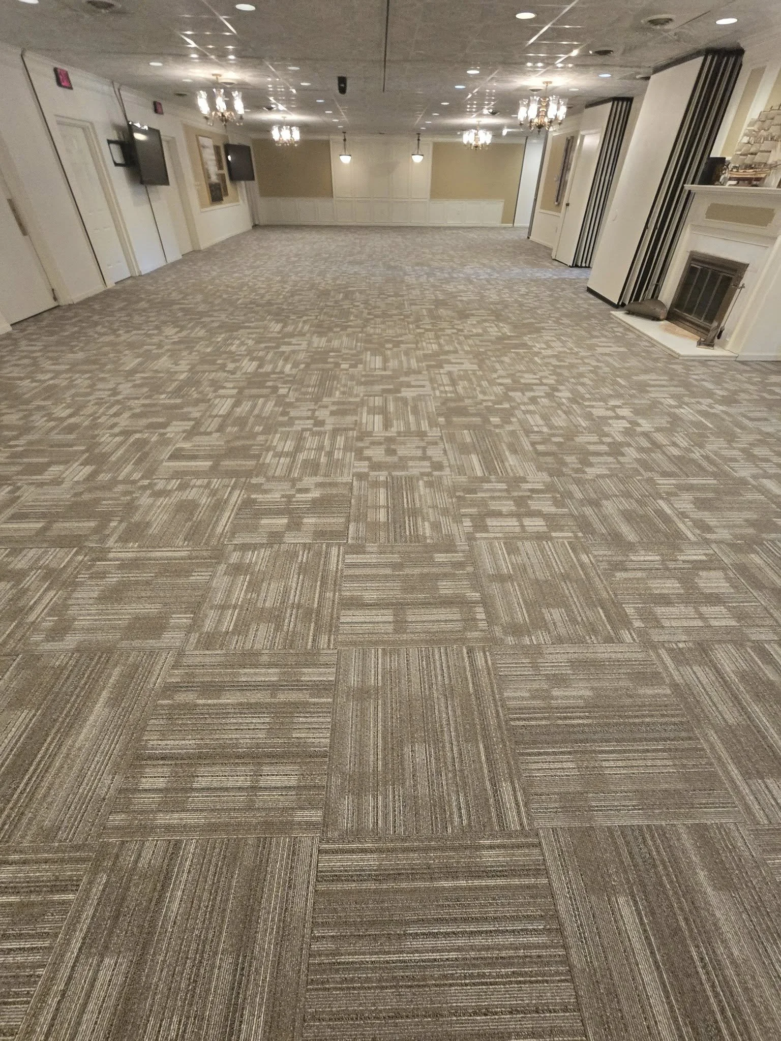 Empty banquet room with patterned carpet, beige walls, multiple chandeliers, and wall-mounted televisions.