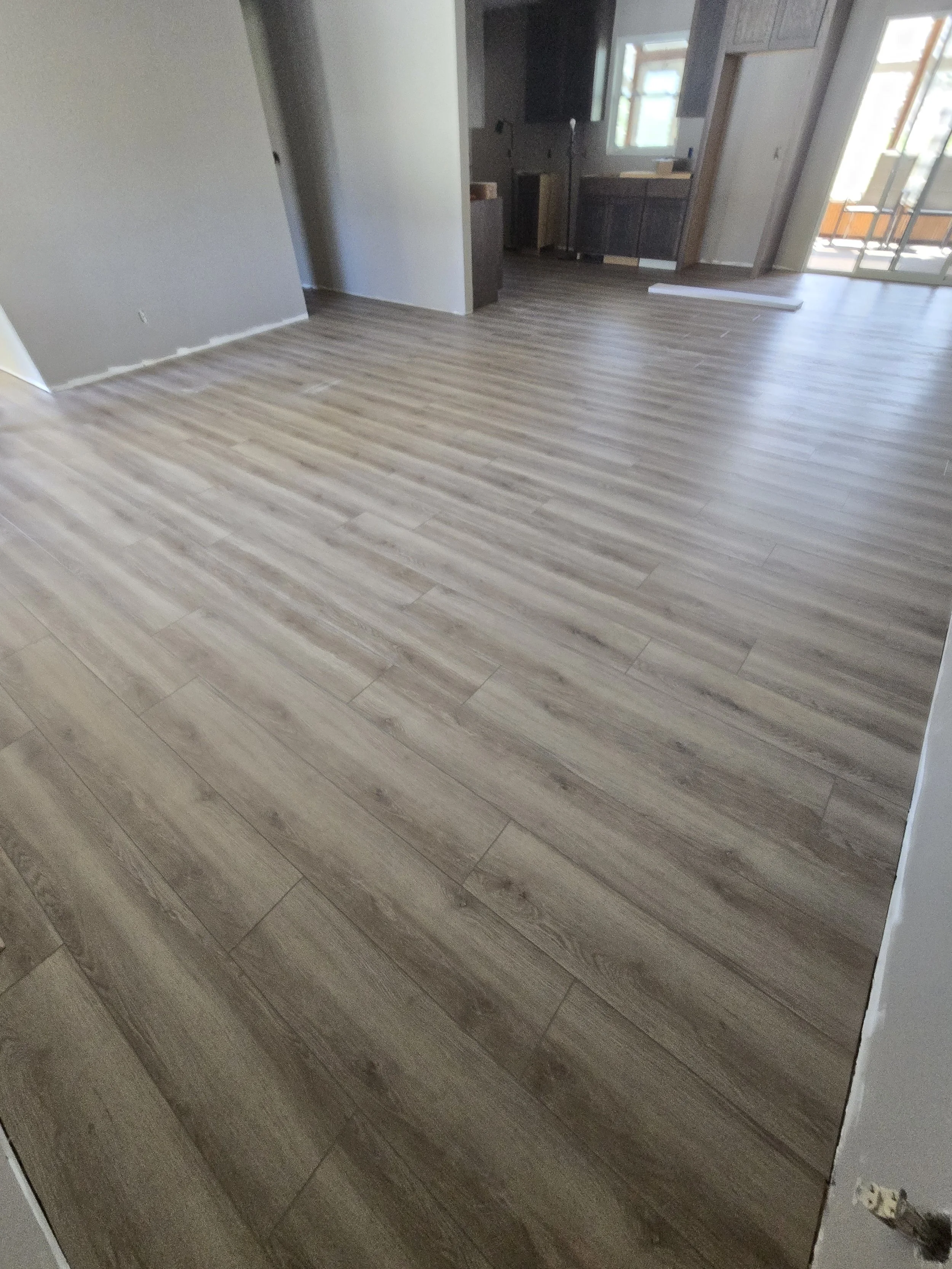 An empty room with wood flooring, part of a kitchen area with dark cabinets and a window, and a sliding glass door leading to an outdoor area.