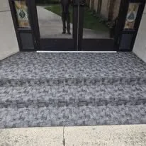 View of a front entrance with grey striped doormat and glass doors, showing a person standing outside reflected in the glass.