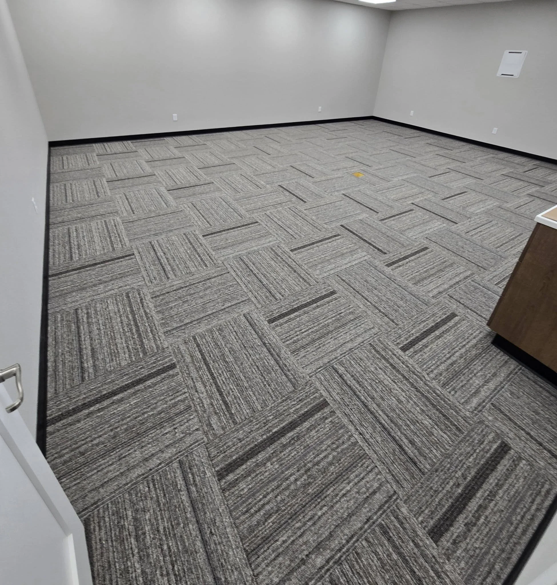 Empty room with gray patterned carpet, white walls, few electrical outlets, and a small vent on the upper right wall.