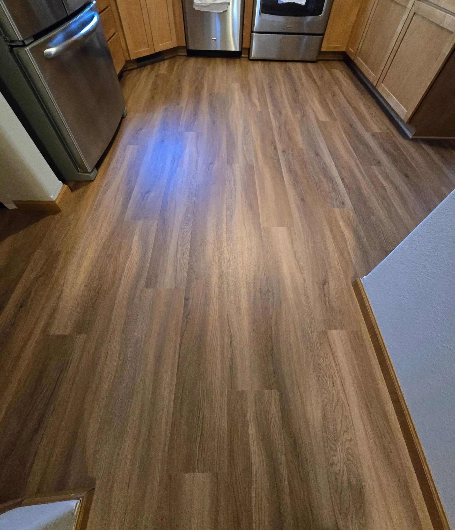 A kitchen with wooden cabinets, stainless steel appliances including a refrigerator, dishwasher, and oven, and a wood floor. The photo is taken from a corner near the wall.