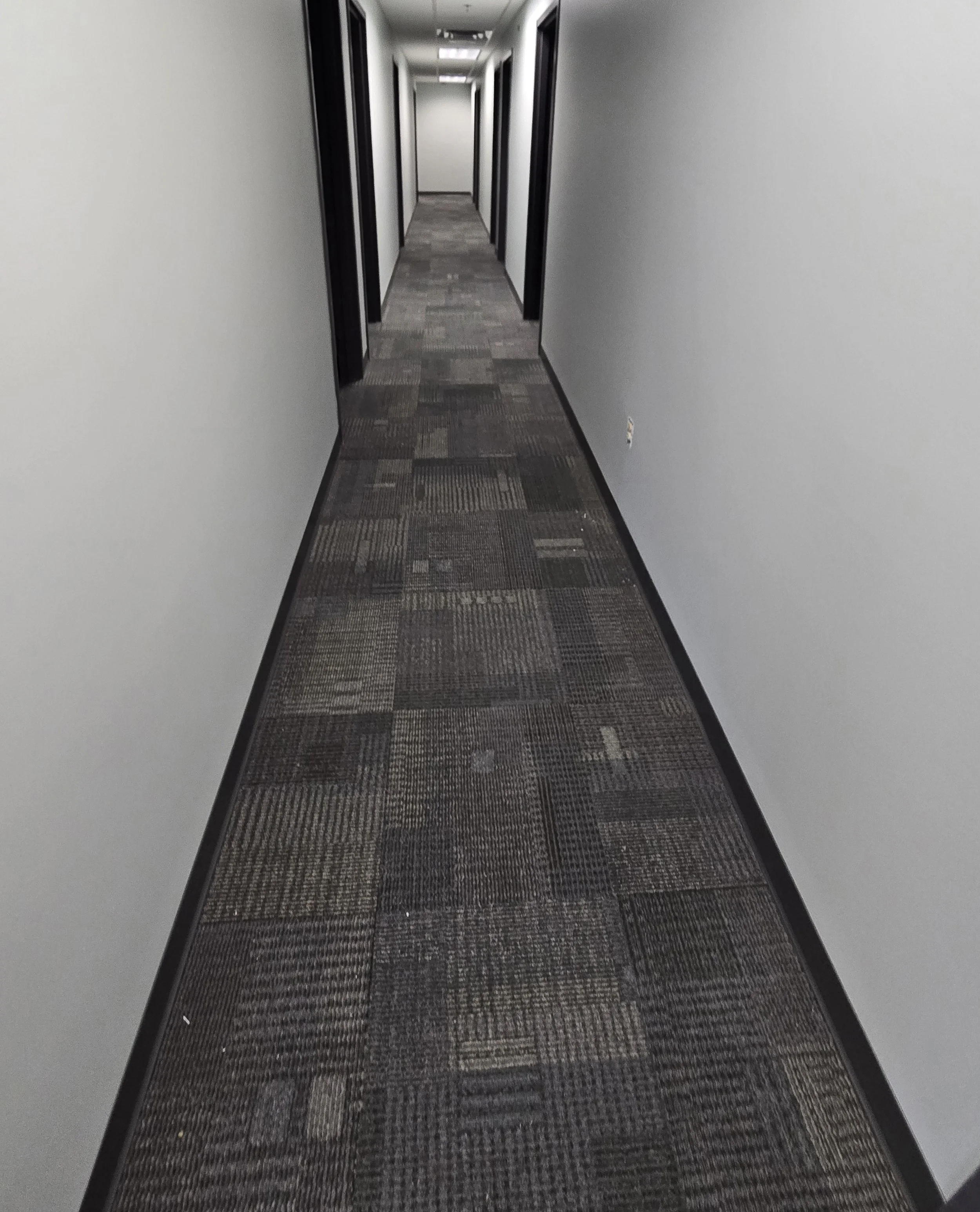 A long, narrow office corridor with a patterned carpet, white walls, black door frames, and ceiling lights, extending to a door at the end.