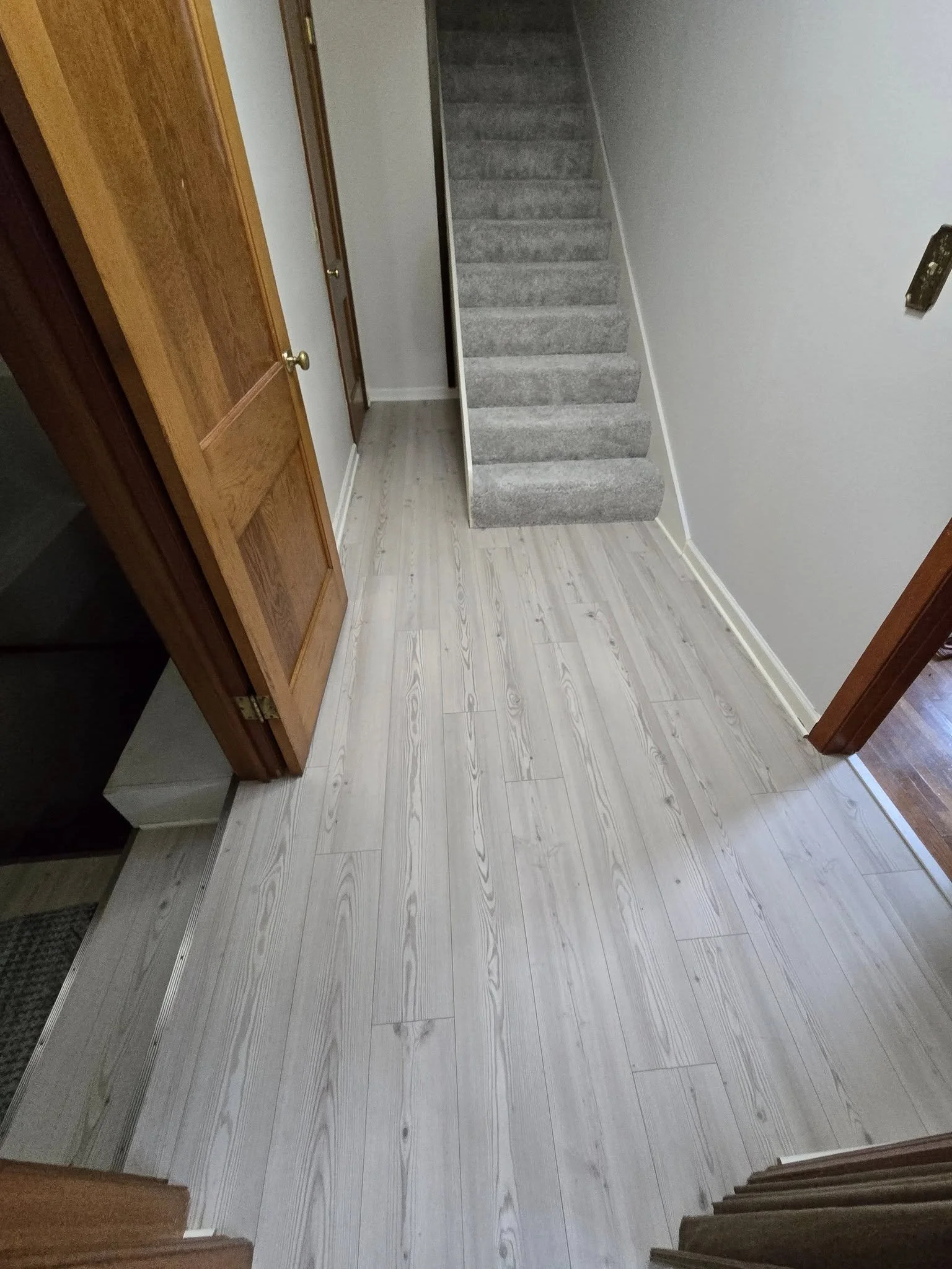 A small hallway with light-colored wood laminate flooring, white walls, and a staircase with gray carpeted steps leading upward. There is a wooden door on the left side and an open doorway on the right. A black object, possibly a remote or device, is
