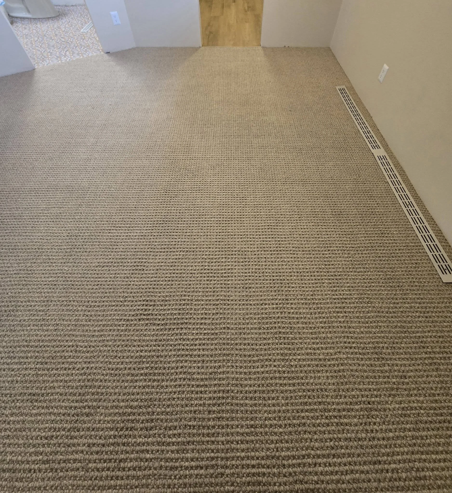 Empty room with beige carpet, a vent on the floor near the right wall, and an electrical outlet on the right wall. A doorway on the left reveals a patterned tile floor.