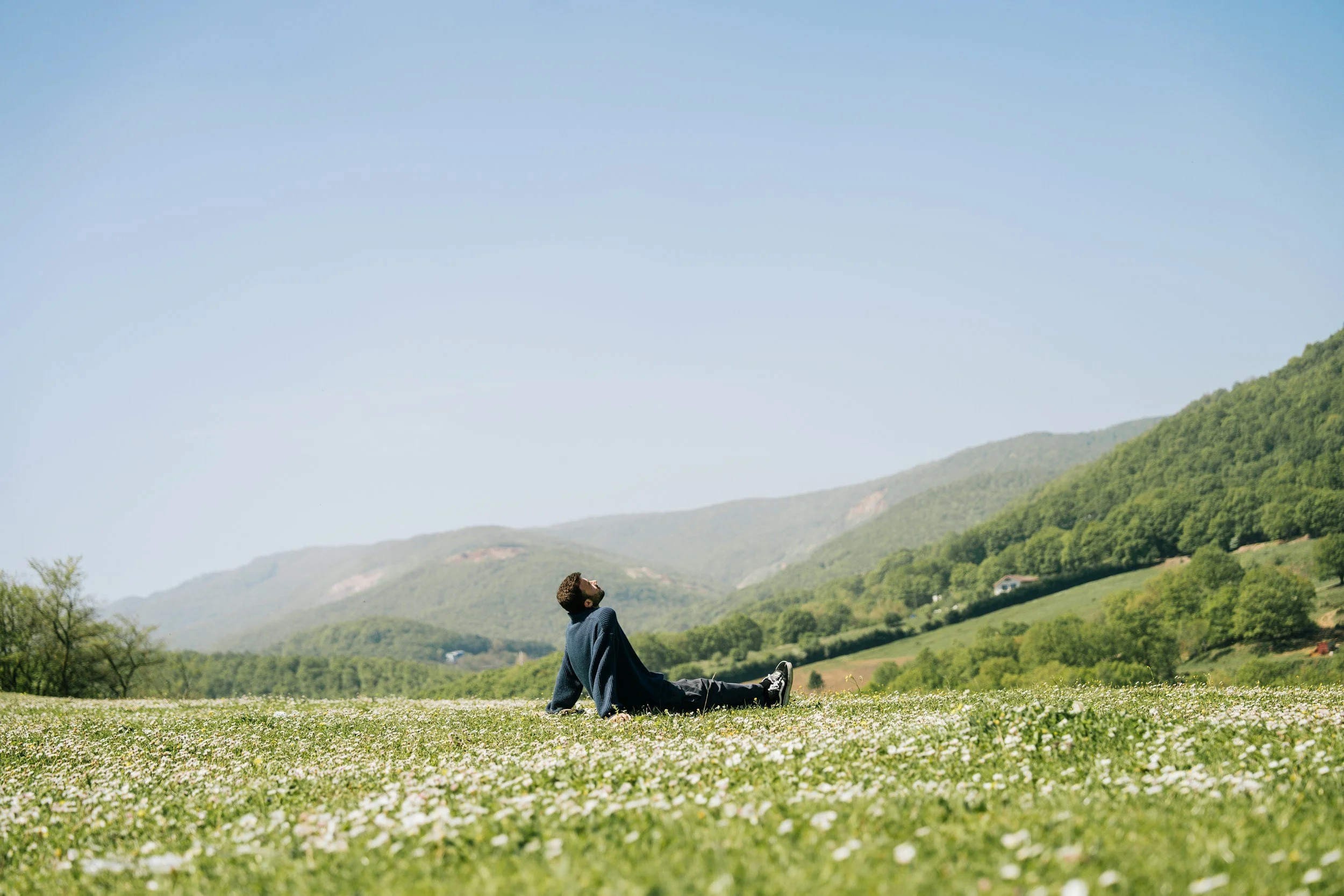 A man with a beard sitting on the grassy field, looking up at the sky with mountains in the background.