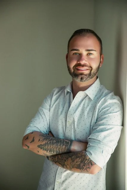 A man with a beard and short hair smiling, wearing a white shirt with rolled-up sleeves and visible tattoos on his forearms, standing against a plain green wall.