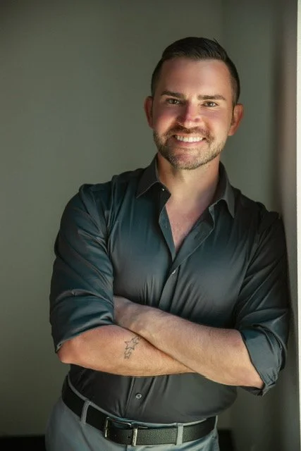 A man with dark hair and a beard in a black dress shirt, smiling and leaning against a wall with arms crossed.