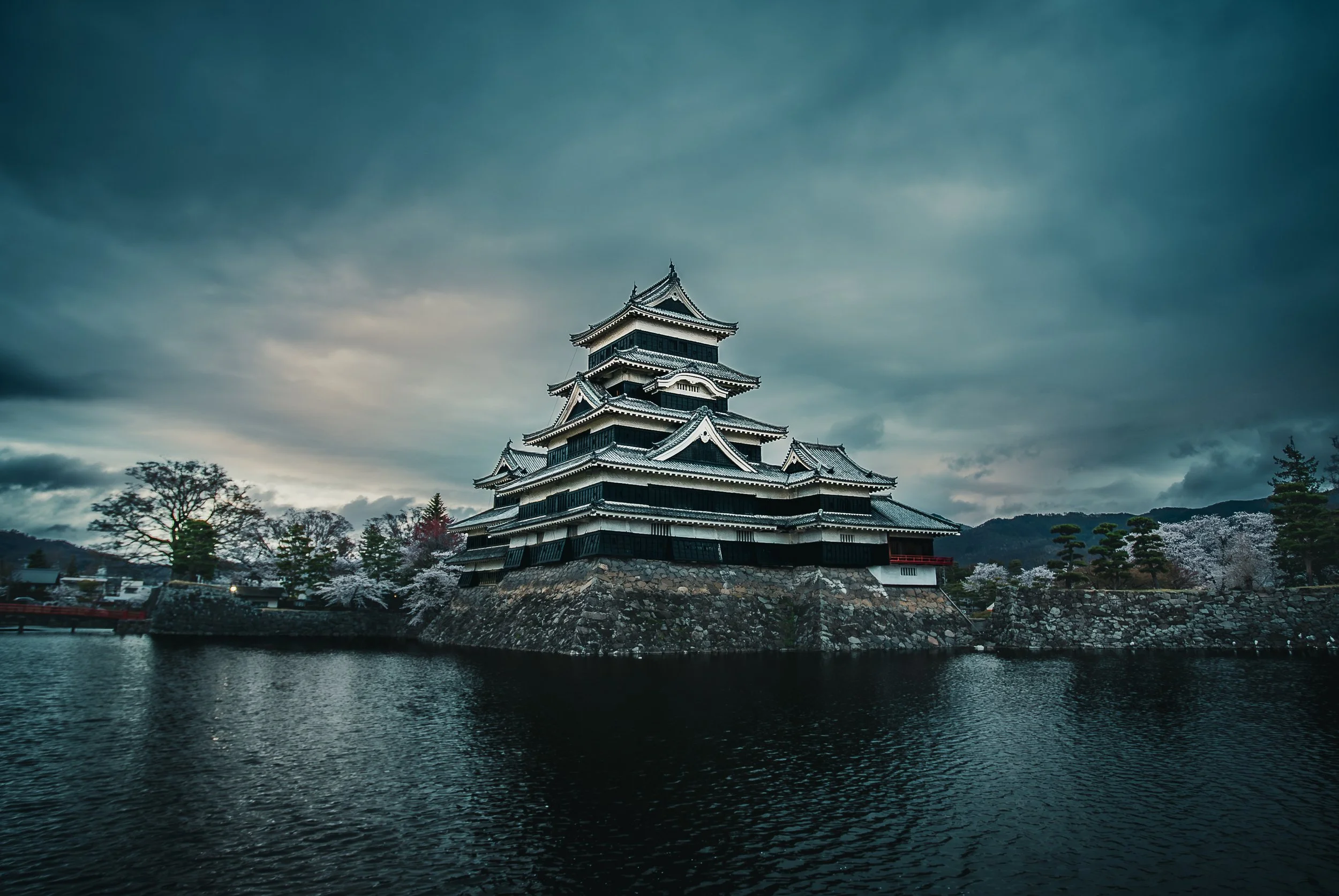 A traditional Japanese castle with multiple tiers and ornate rooftops, surrounded by a moat and trees with cherry blossoms, under a cloudy sky.