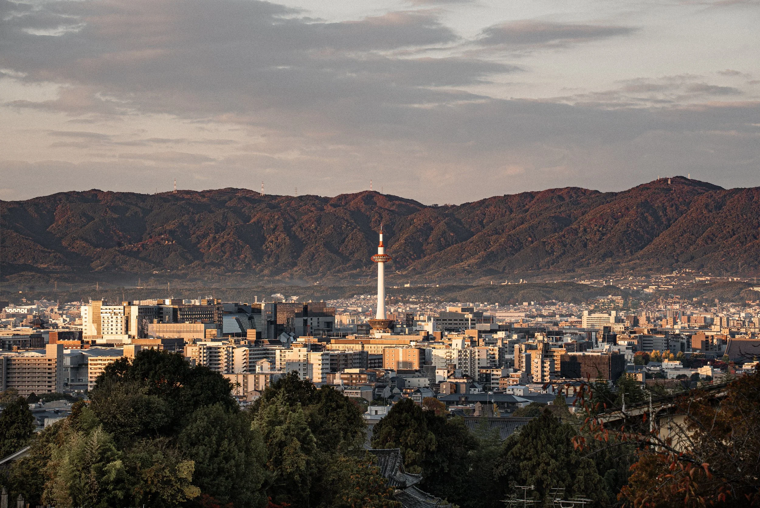City skyline with buildings and a prominent tower, set against a mountainous backdrop under a cloudy sky at sunset.