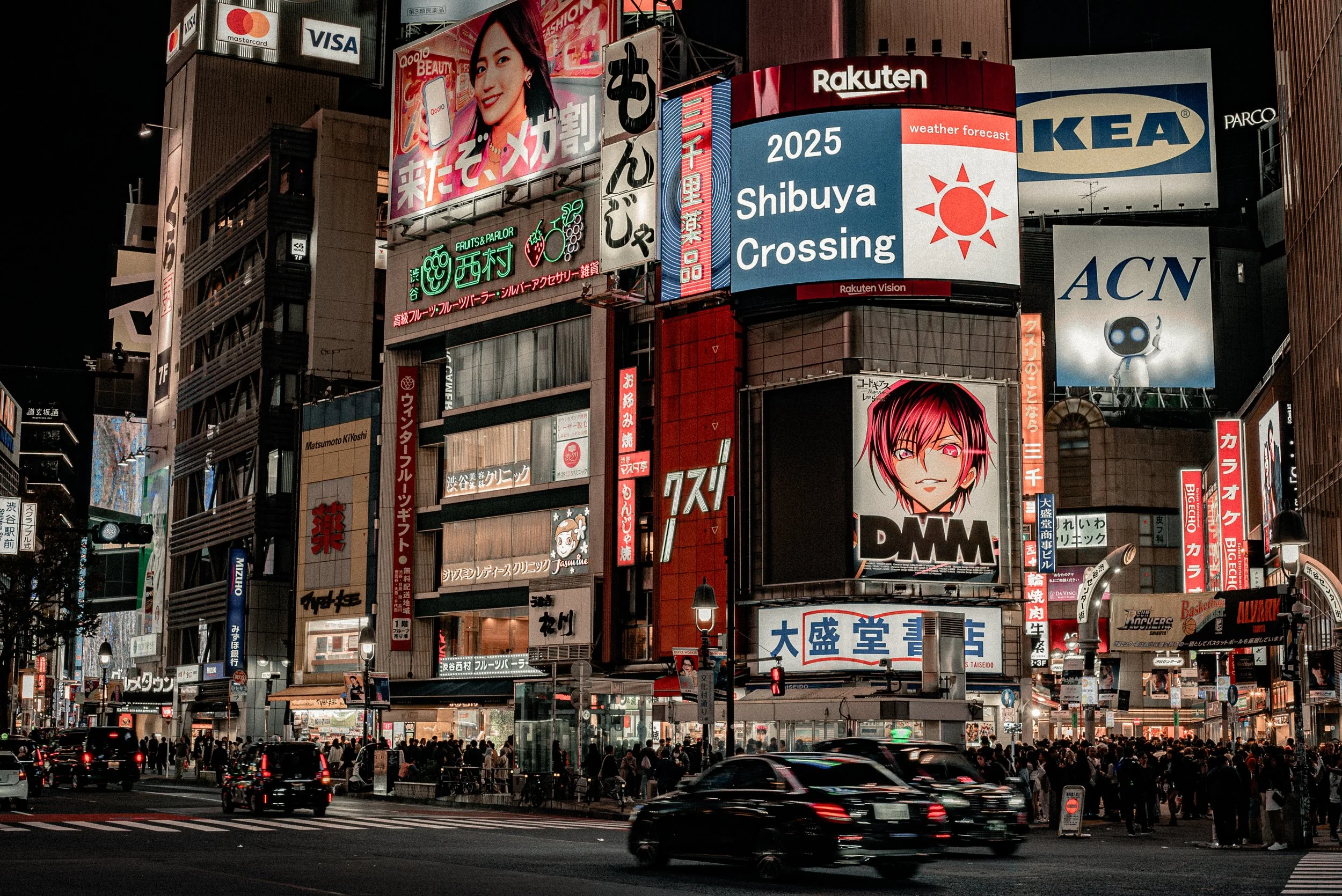 A nighttime city scene with illuminated billboards and advertisements on tall buildings, busy traffic with cars, and many pedestrians on the street.