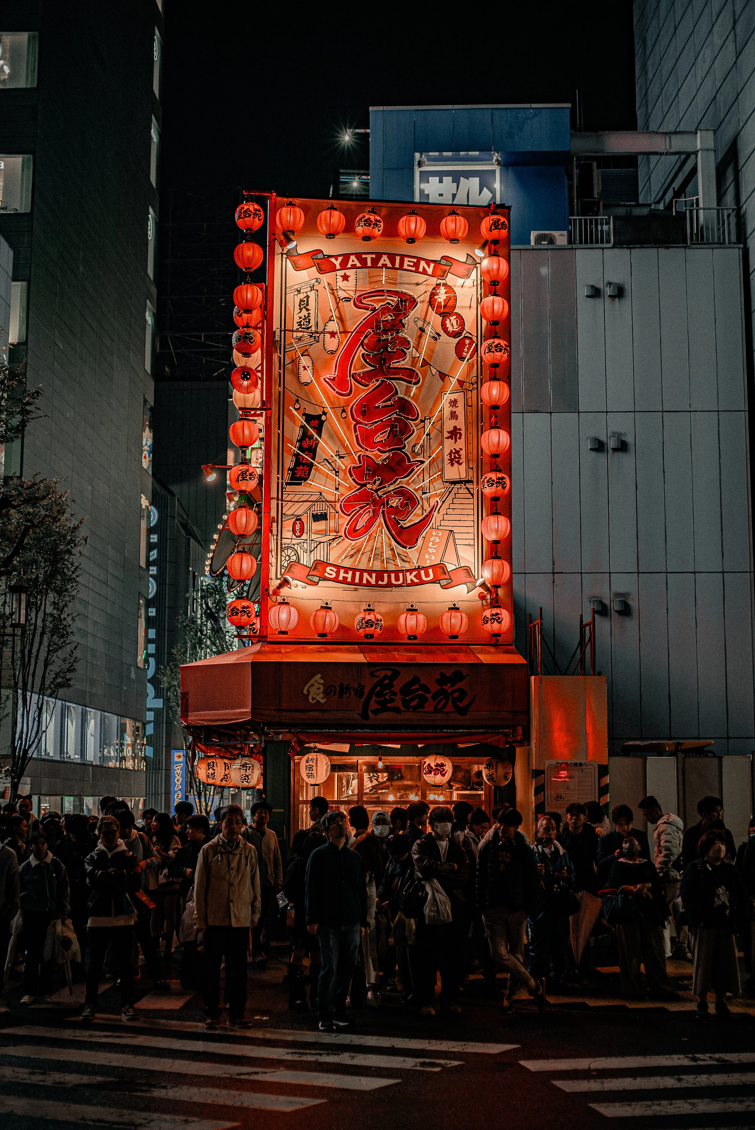 Night scene of a busy street in Shinjuku, Japan, with a bright illuminated sign featuring Japanese characters and red paper lanterns, and a crowd of people walking in front of it.