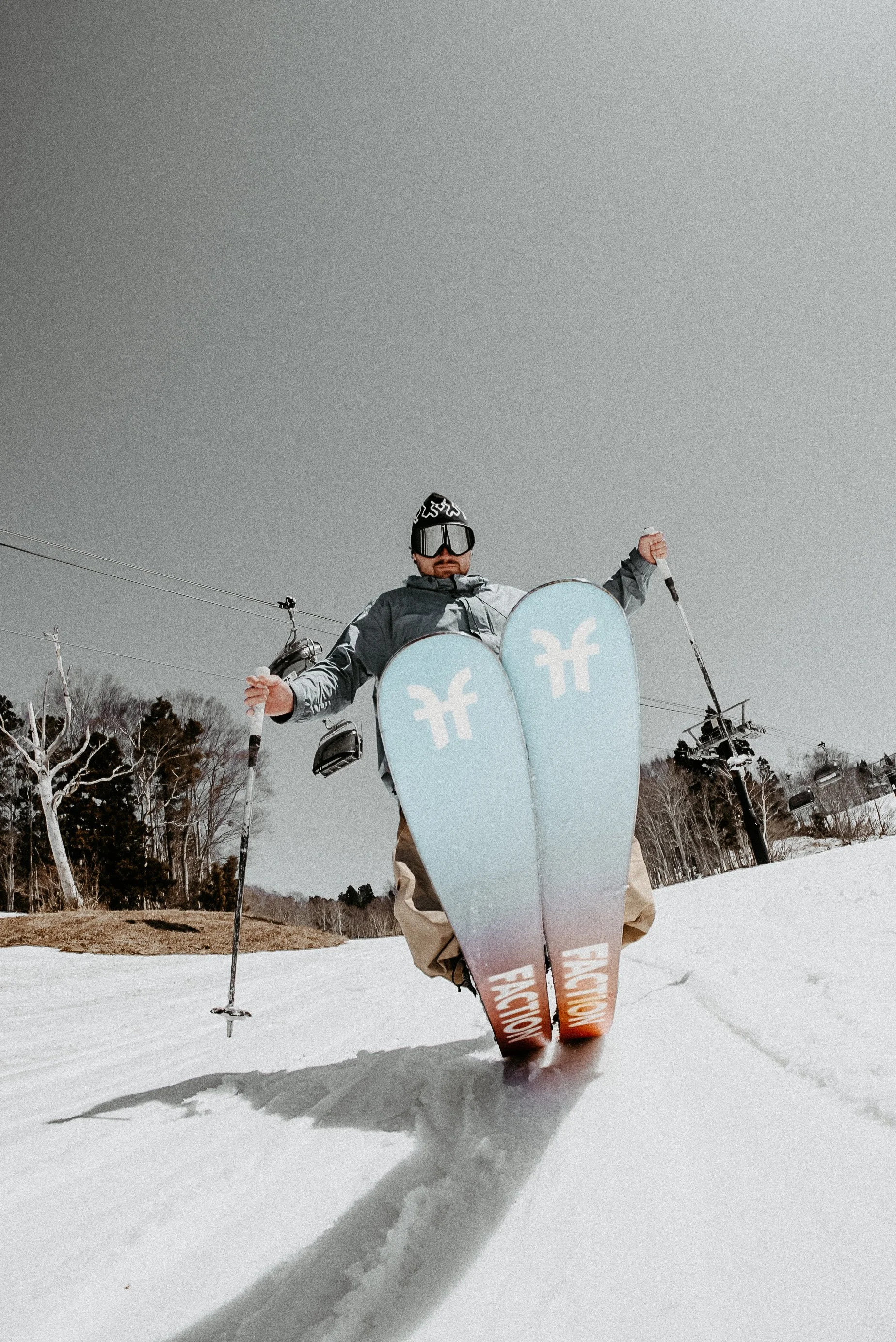 Skier wearing goggles, a jacket, and ski pants standing in snow on a ski slope, holding ski poles, with ski boots and skis with branding, surrounded by trees and ski lifts in the background.