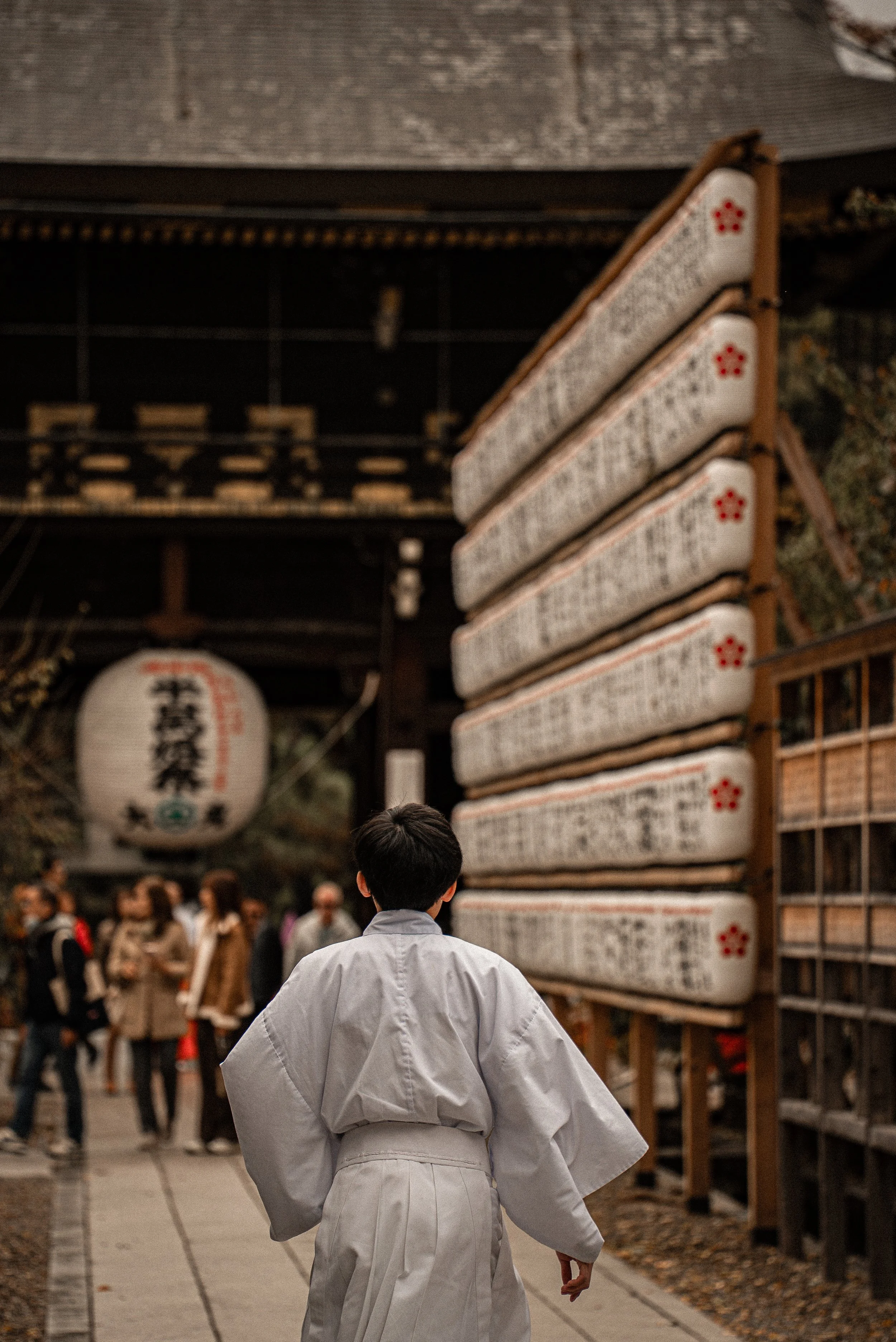 A person dressed in traditional white martial arts attire walking through a busy outdoor area with Japanese lanterns and horizontal wooden plaques hanging along a wall in the background, possibly at a shrine or temple.