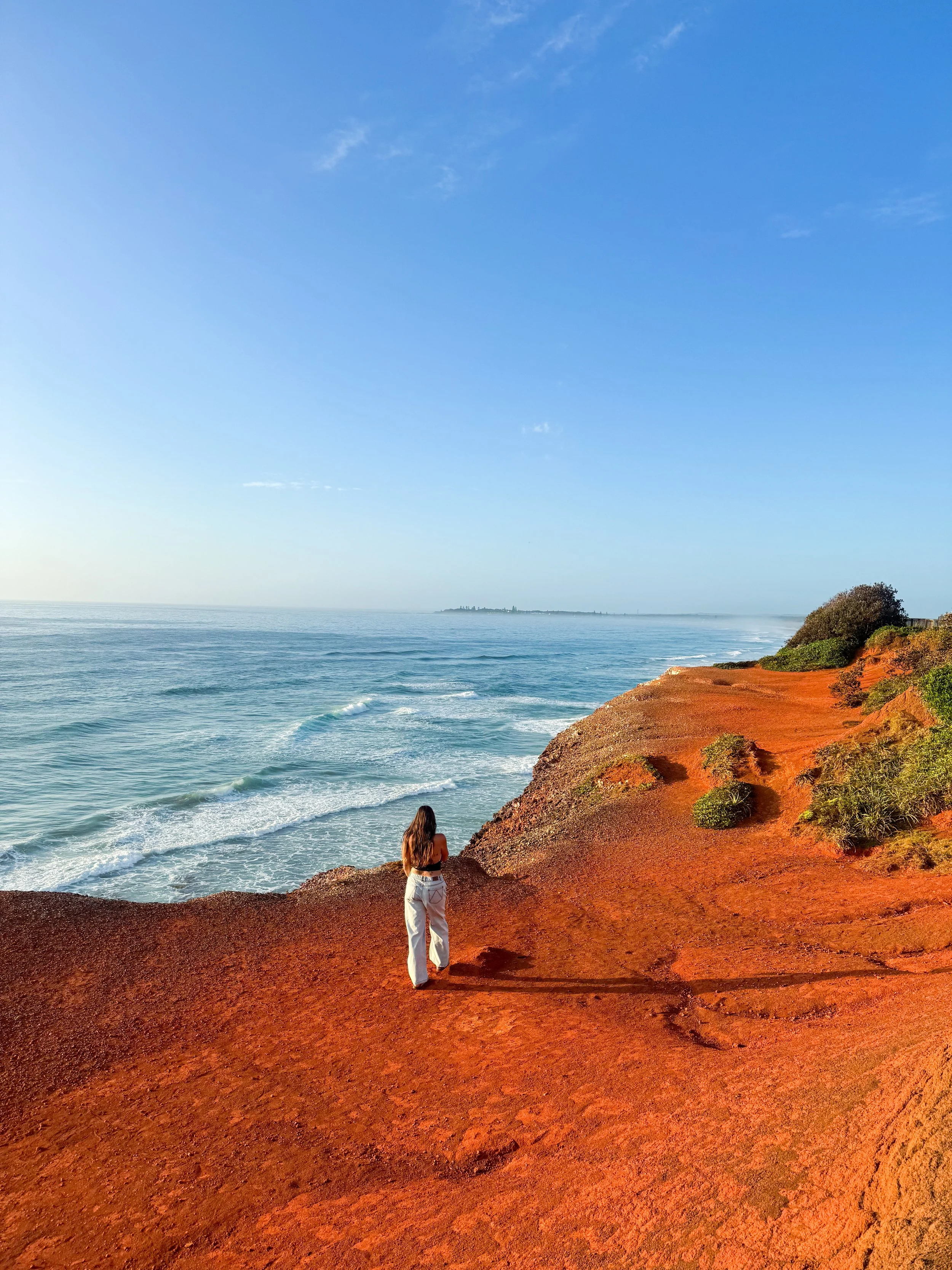 A woman standing on a red rocky cliff overlooking the ocean with waves crashing below. The sky is blue with some clouds, and there is a distant landmass on the horizon. Vegetation is present on the cliff edge.